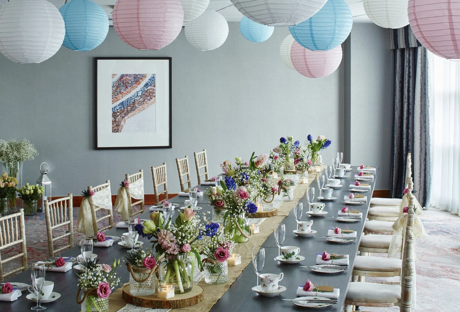 A long dining table decorated for a celebration with floral centerpieces, tea cups, and glasses; paper lanterns in pink, white, and blue hanging from the ceiling; a framed abstract artwork on the wall; curtains and a window in the background.