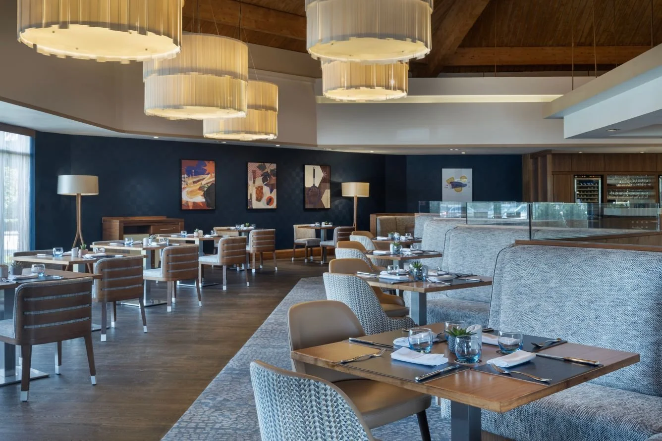 Interior of a modern restaurant with wooden floors, various tables set with glassware and utensils, comfortable seating, abstract artwork on navy blue walls, tall lamps, and large drum-shaped pendant lights hanging from the ceiling.