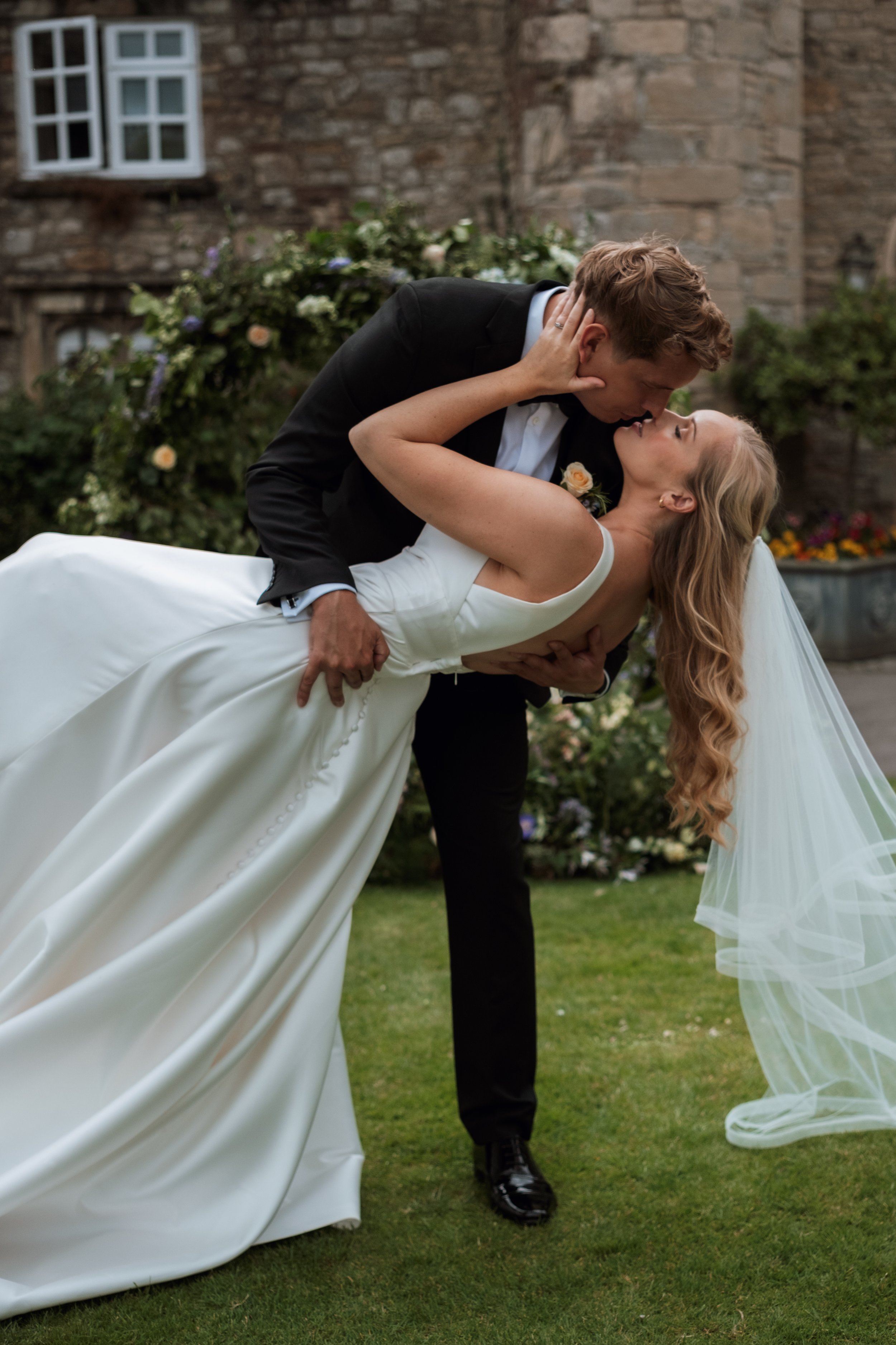 A bride and groom are dancing outside, with the groom dipping the bride backward while holding her around the waist. They are passionately close, with the groom leaning in for a kiss and the bride gazing up at him. The scene is set against a rustic brick building with floral decorations in the background.