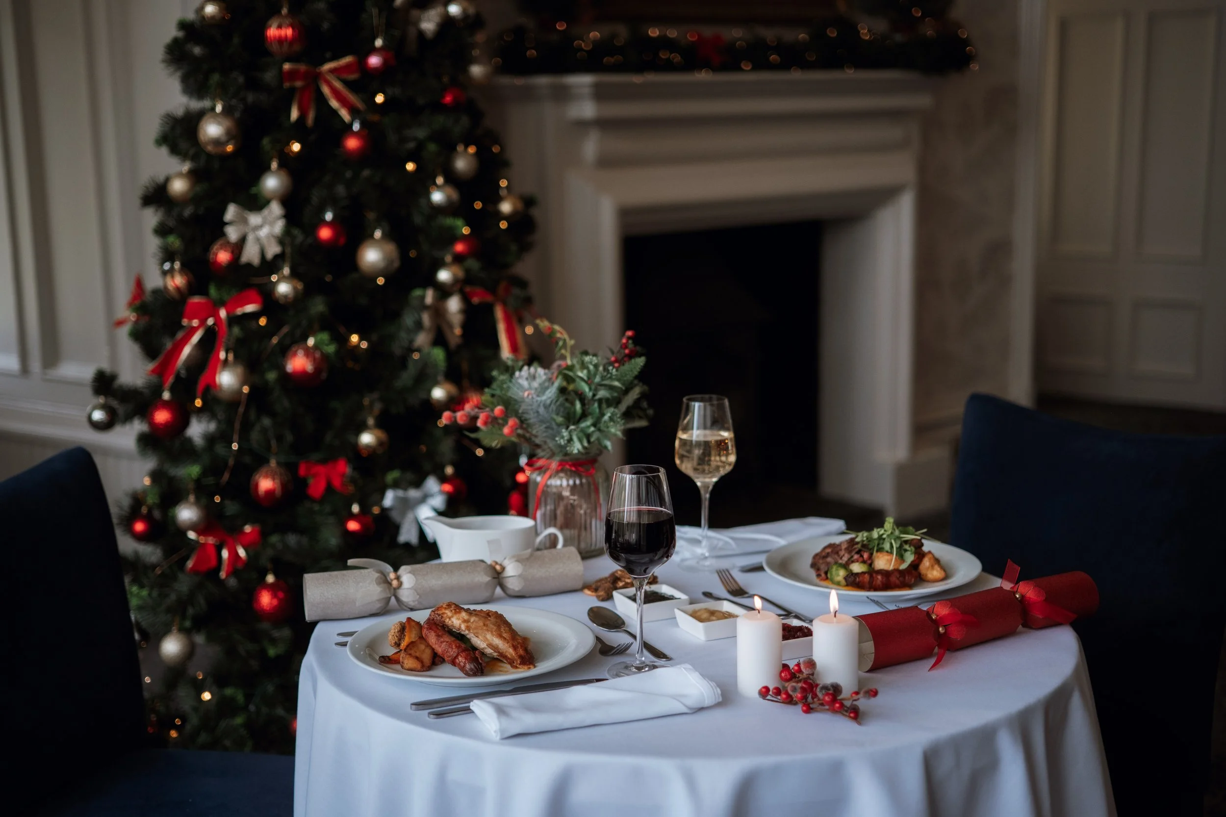 A festive dinner table set with white tablecloth, candles, and holiday decorations. In the background, a decorated Christmas tree with red and silver ornaments and ribbons. The table has plates of food, glasses of drinks, and holiday-themed decor.