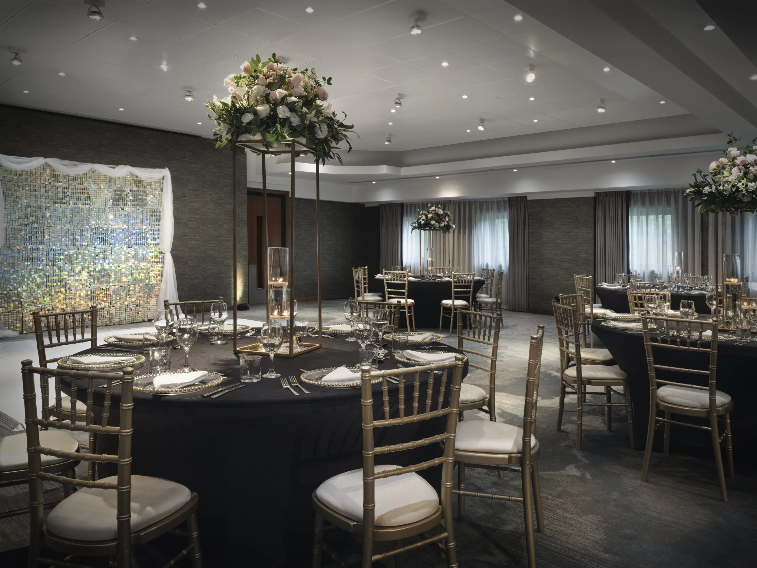 Elegant banquet hall decorated with tall floral arrangements of pink and white flowers on wooden stands, set for a formal event with round tables covered in black tablecloths, gold chairs, and glassware. The room has a high ceiling with recessed lighting, sheer gray curtains, and a shimmering backdrop on one wall.