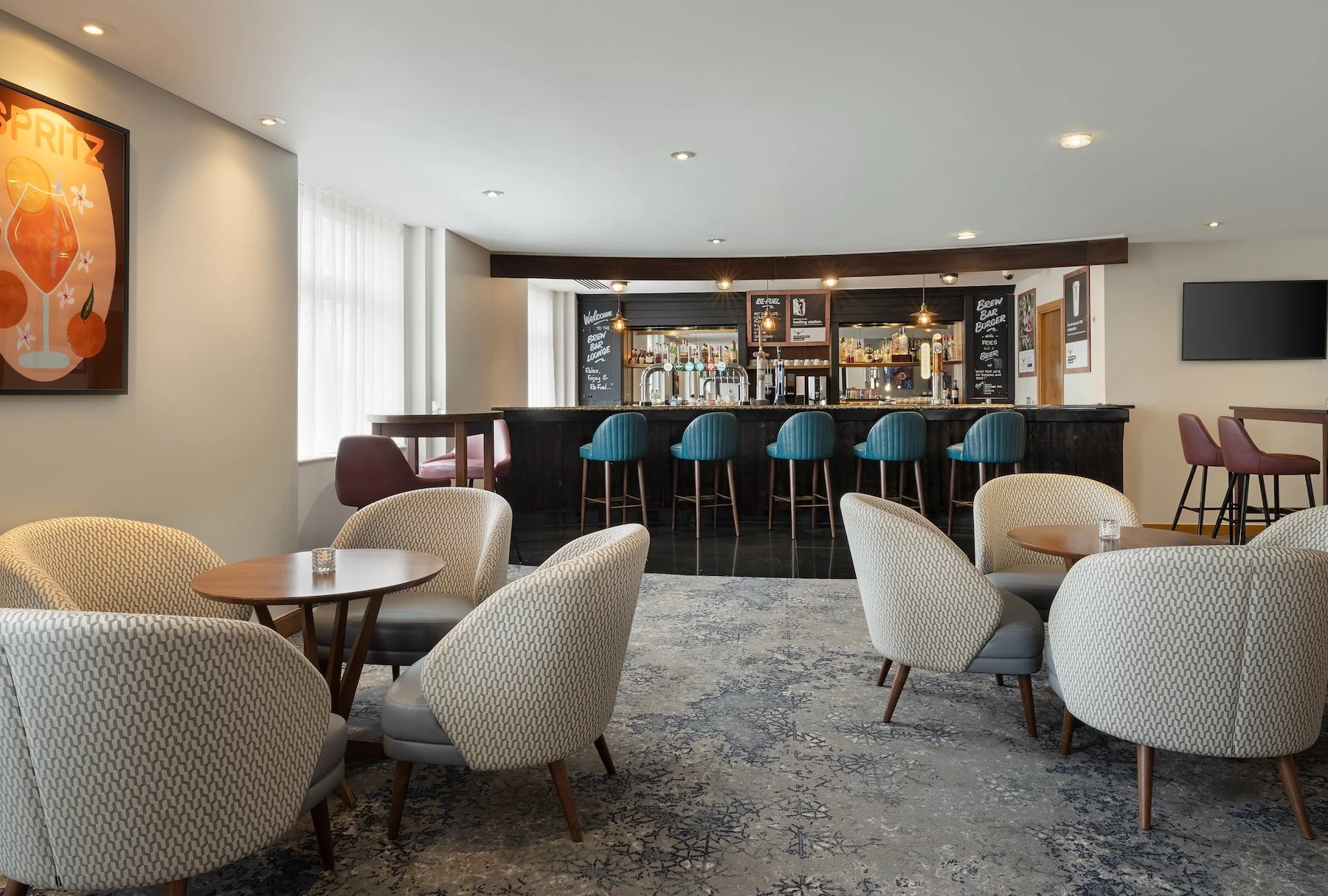 Interior of a modern bar with seating area, including several patterned armchairs around small wooden tables, and a bar with blue cushioned stools. The bar area has various bottles and a chalkboard menu, with bright lighting and a large window with vertical blinds.