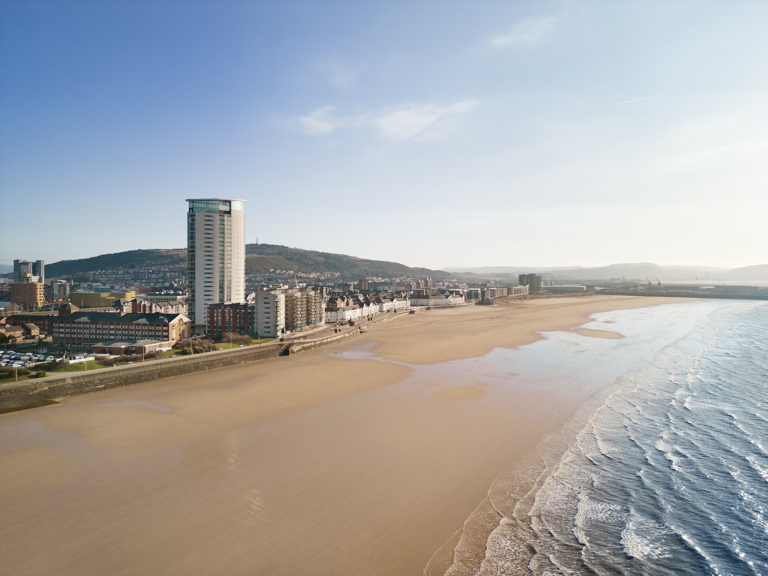 A cityscape along a beach with tall buildings, a sandy shoreline, and calm ocean water under a clear blue sky.
