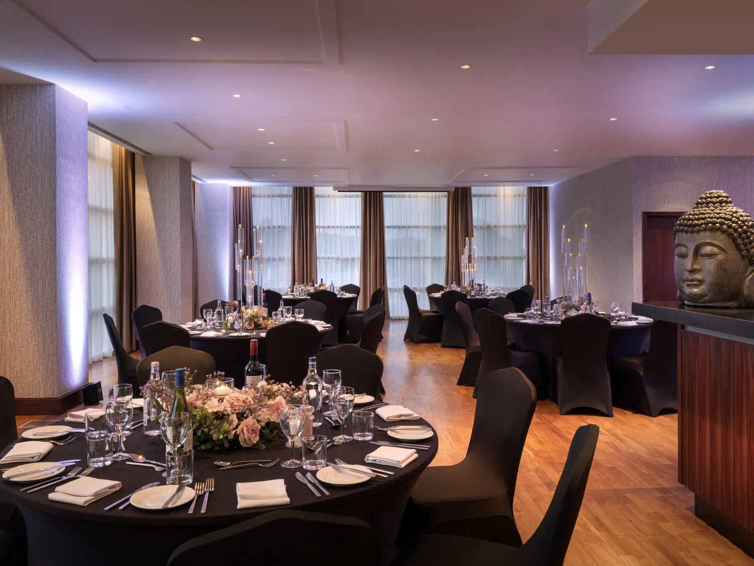 Elegant banquet room with round tables set for dining, decorated with centerpieces, wine bottles, and glassware, featuring large windows with drapes and a large Buddha statue.