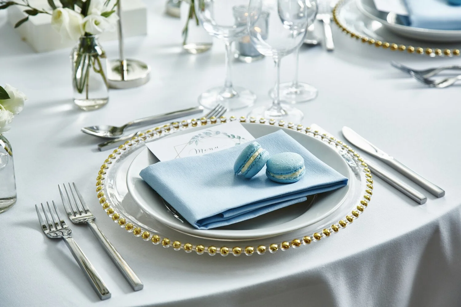 Elegant table setting with a white tablecloth, a gold beaded charger plate, a blue cloth napkin with two blue macarons on top, and silverware including a fork, knife, and spoon. There are wine glasses and small vases with white flowers in the background.