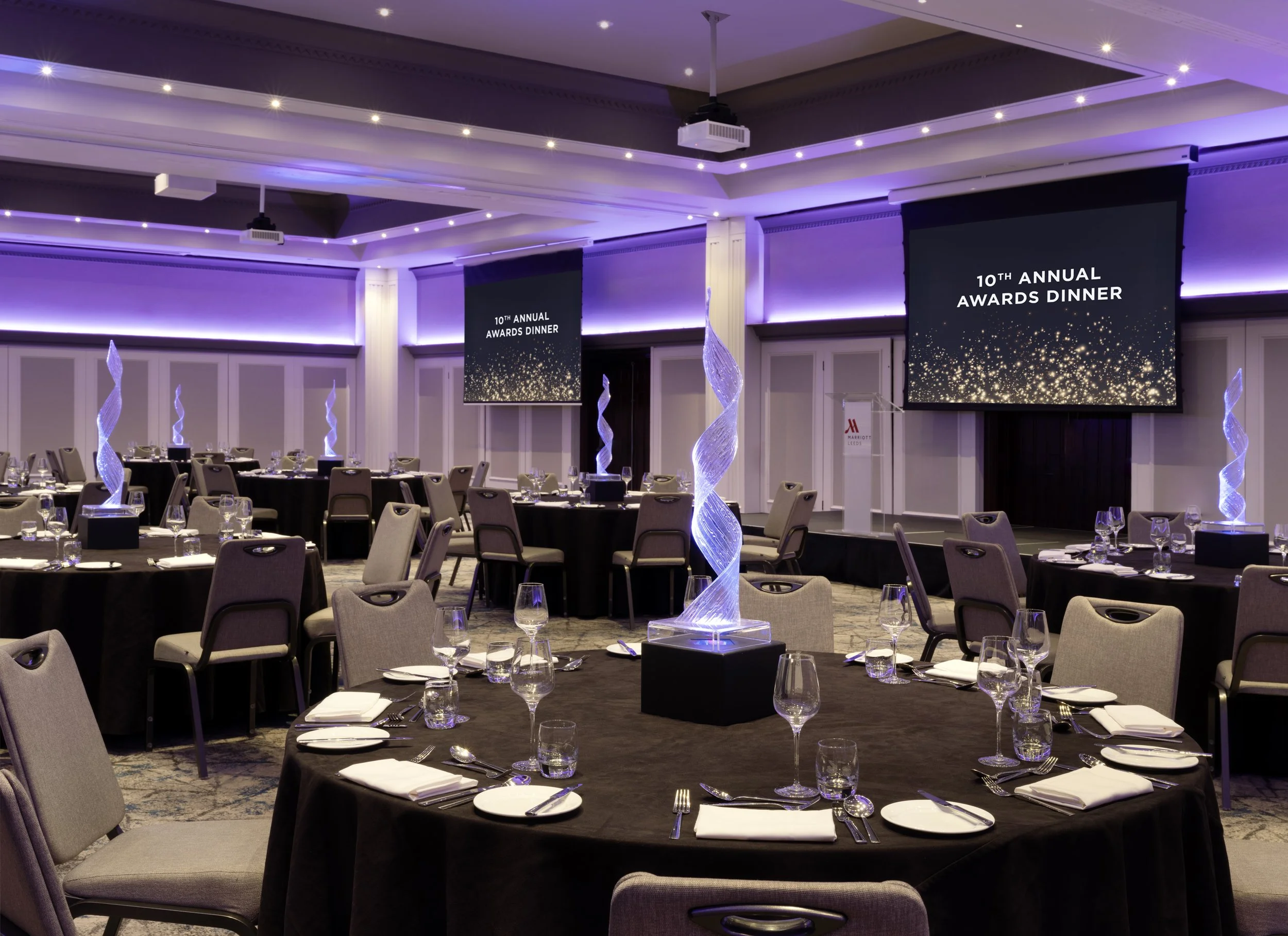 Elegant banquet hall decorated for the 10th Annual Awards Dinner with round tables set with silverware, glasses, and white napkins, illuminated by purple lighting and modern spiral centerpieces.