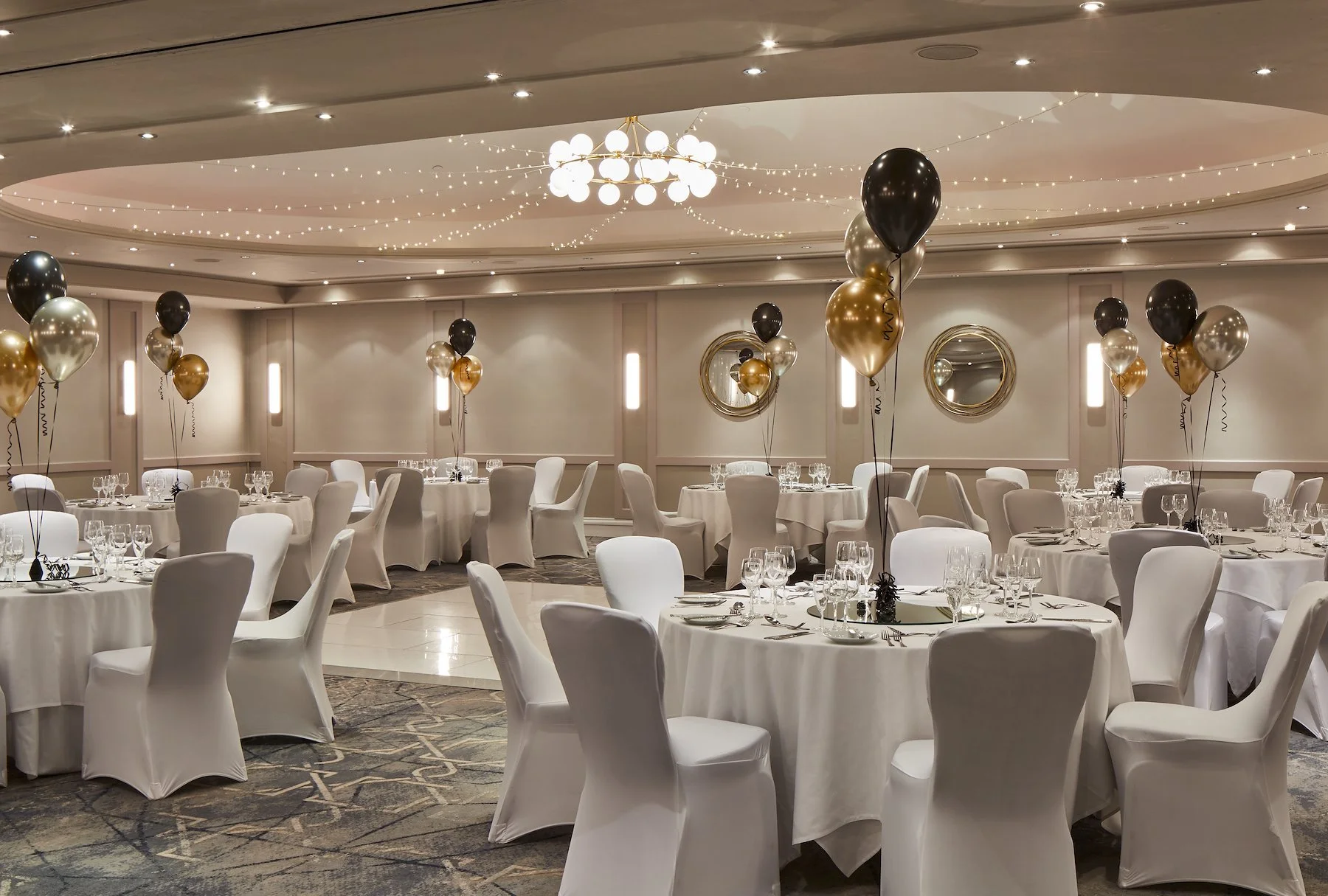 Elegant banquet hall decorated with white tablecloths, white chairs, black, gold, and silver balloons, and string lights on the ceiling, set for a celebration or wedding reception.
