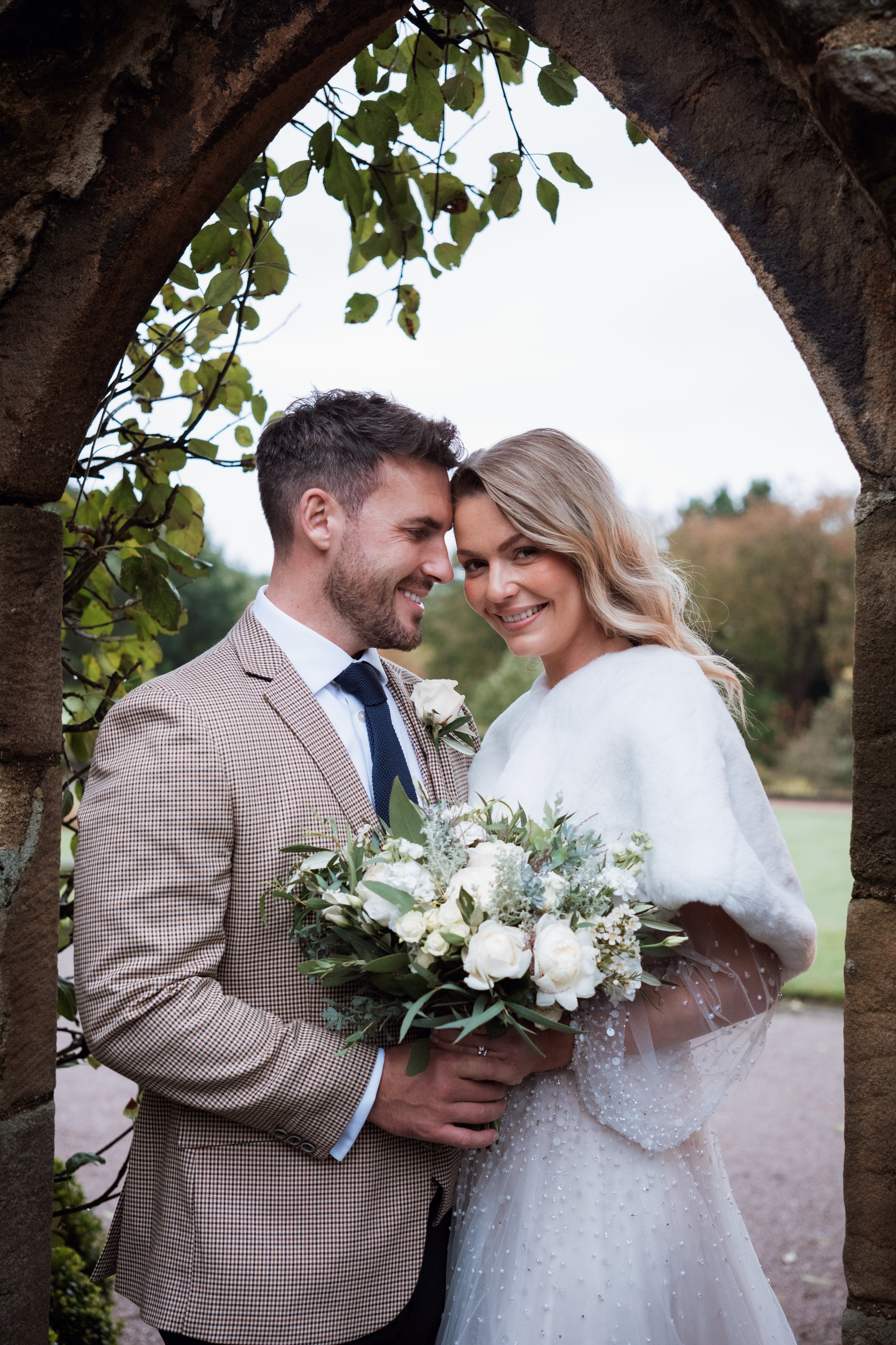 A smiling bride holding a bouquet of white flowers and greenery, standing with a groom in front of a stone archway outdoors. The groom is wearing a checkered suit, white shirt, and dark tie, and the bride is wearing a white dress with pearl embellishments and a white shawl.