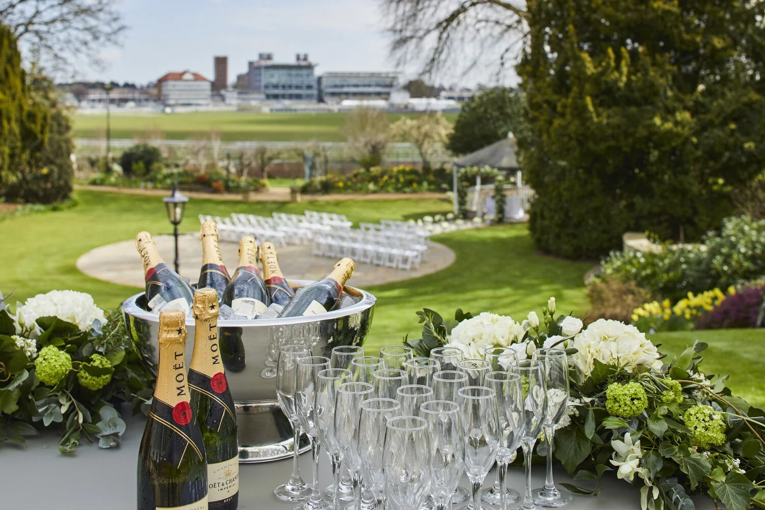 Champagne bottles in ice bucket, empty glasses, and floral decorations on outdoor table, with a garden and lawn in the background.