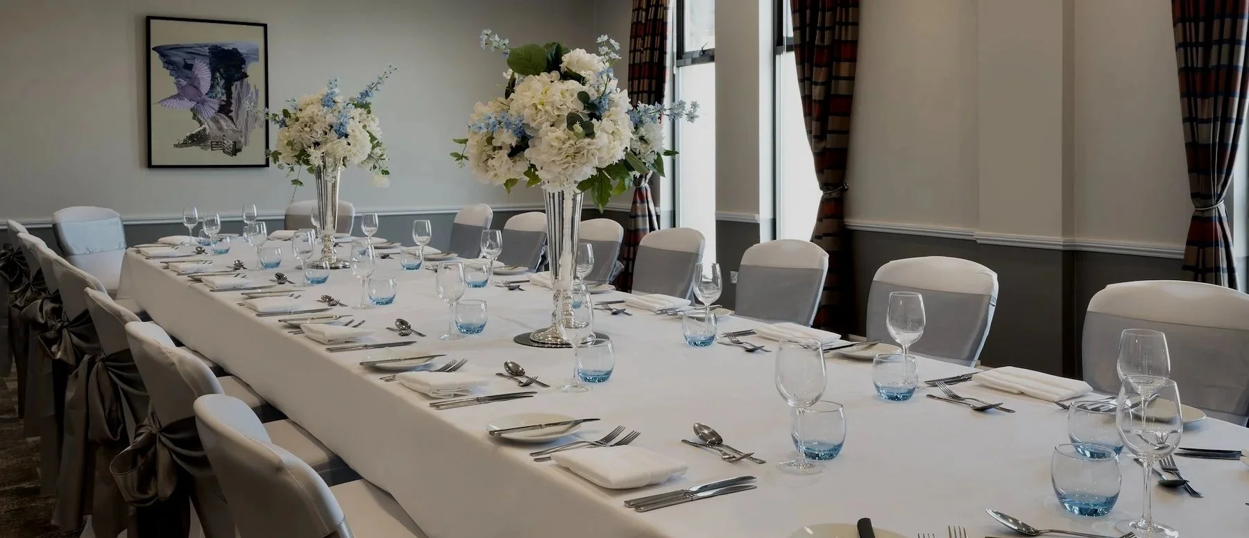 Elegant dining table set for a formal event with white tablecloths, tall floral centerpieces, wine glasses, water glasses, and neatly arranged silverware in a decorated room with large windows and framed artwork.
