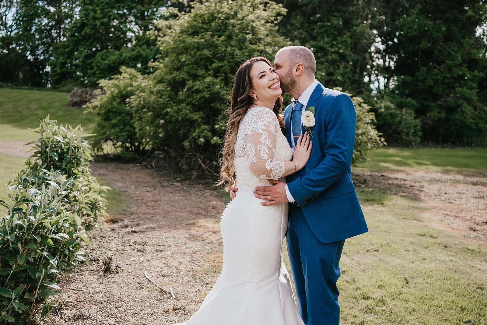 A newlywed couple sharing a kiss and laugh outdoors in a garden setting, with greenery and trees in the background.