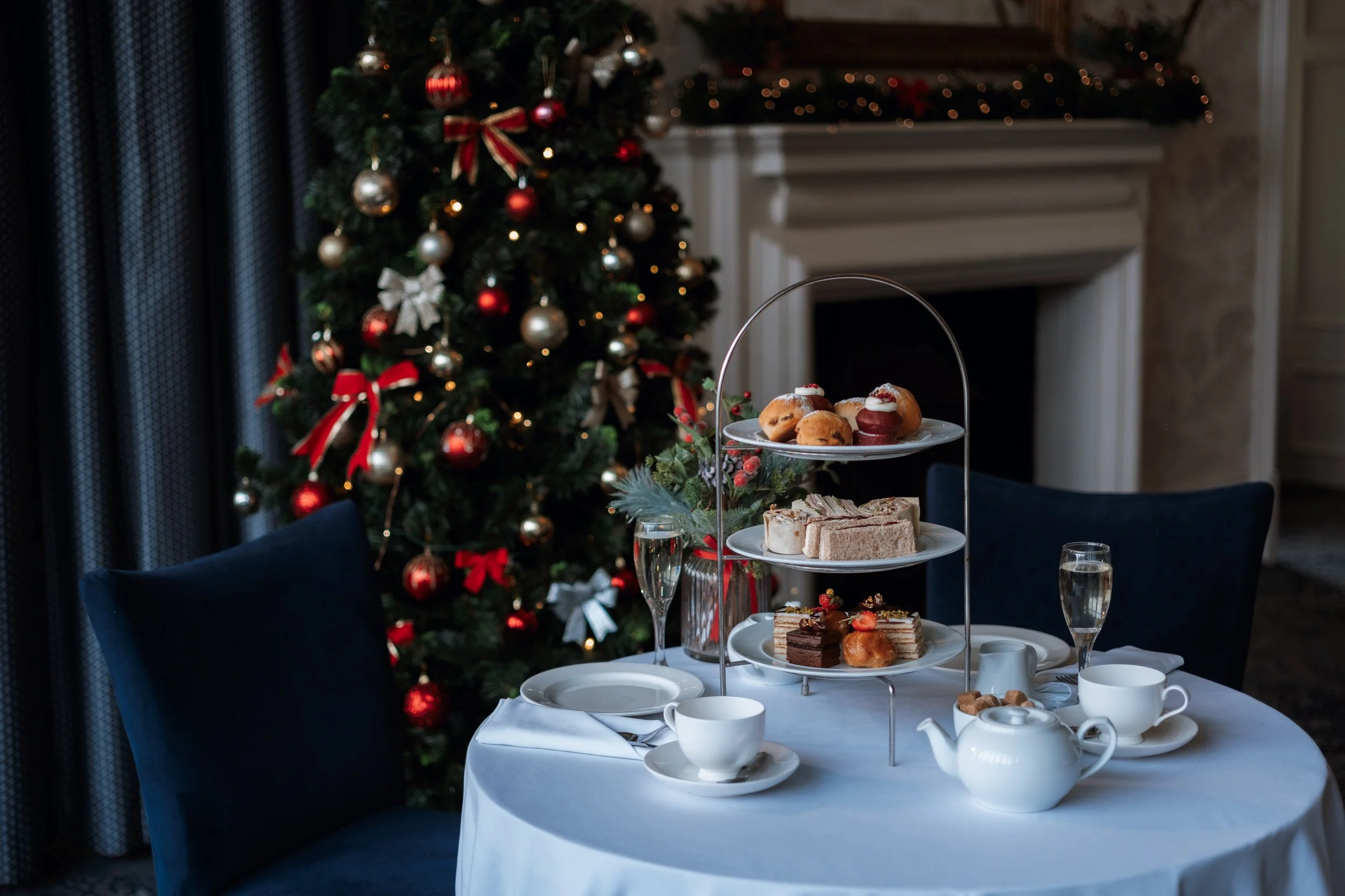 A Christmas-themed table setting featuring a three-tiered stand filled with assorted desserts, including cakes and pastries, with a decorated Christmas tree with red and gold ornaments in the background.