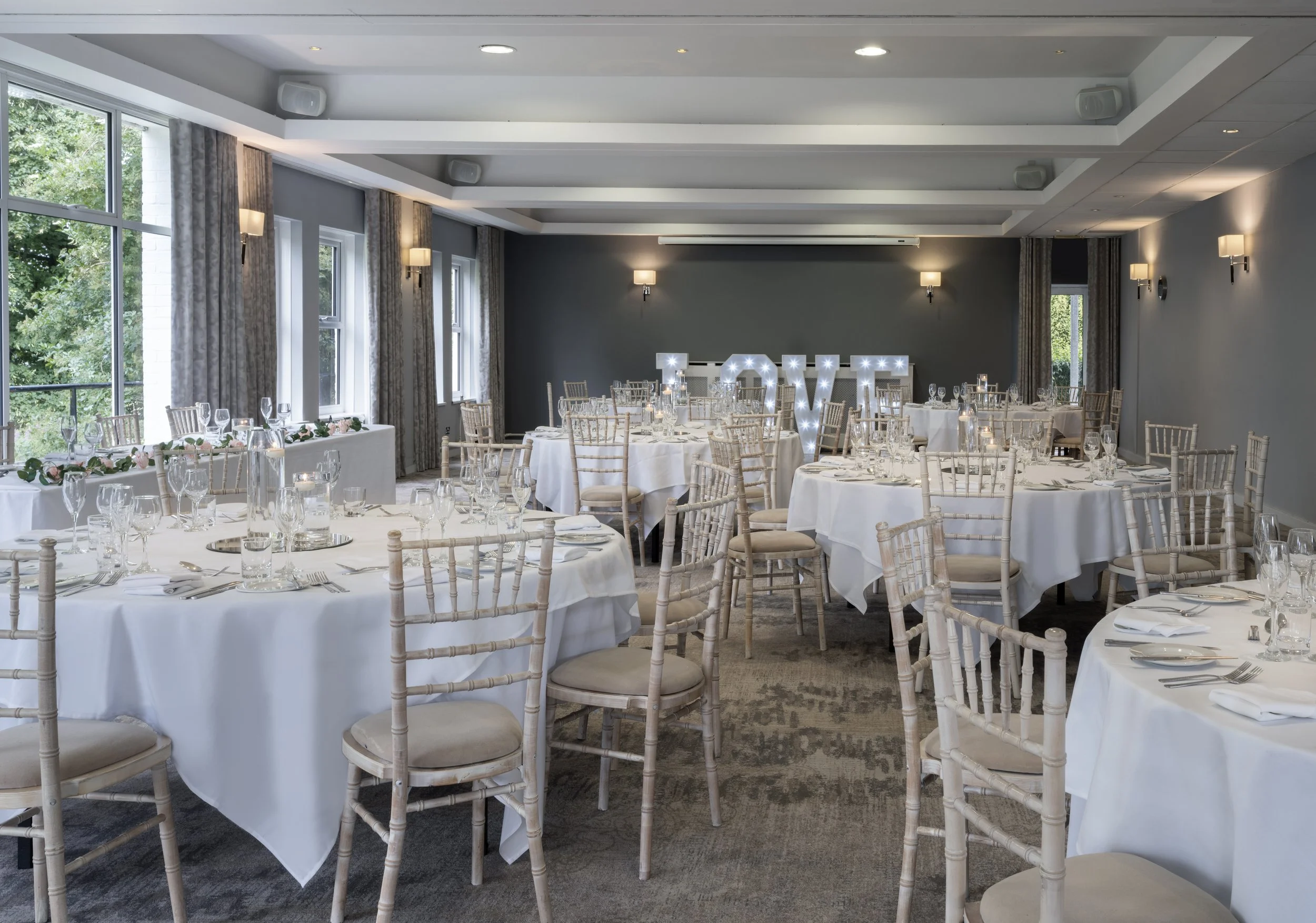 Banquet hall decorated for a wedding reception with round tables, white tablecloths, and elegant chairs. A large sign spelling 'LOVE' with lights is at the back of the room.