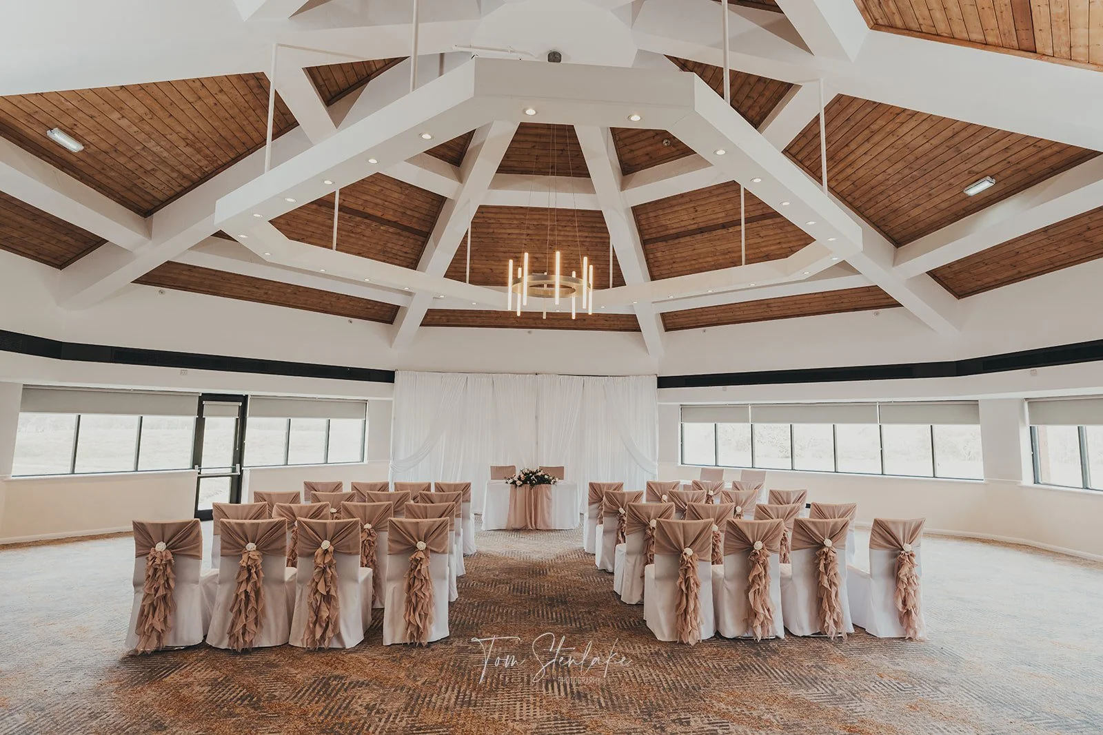 Elegant wedding ceremony space with round tables covered in white cloth and decorated with beige bows, facing a head table with floral arrangements, under a large wooden and white ceiling with a gold modern chandelier, surrounded by windows.
