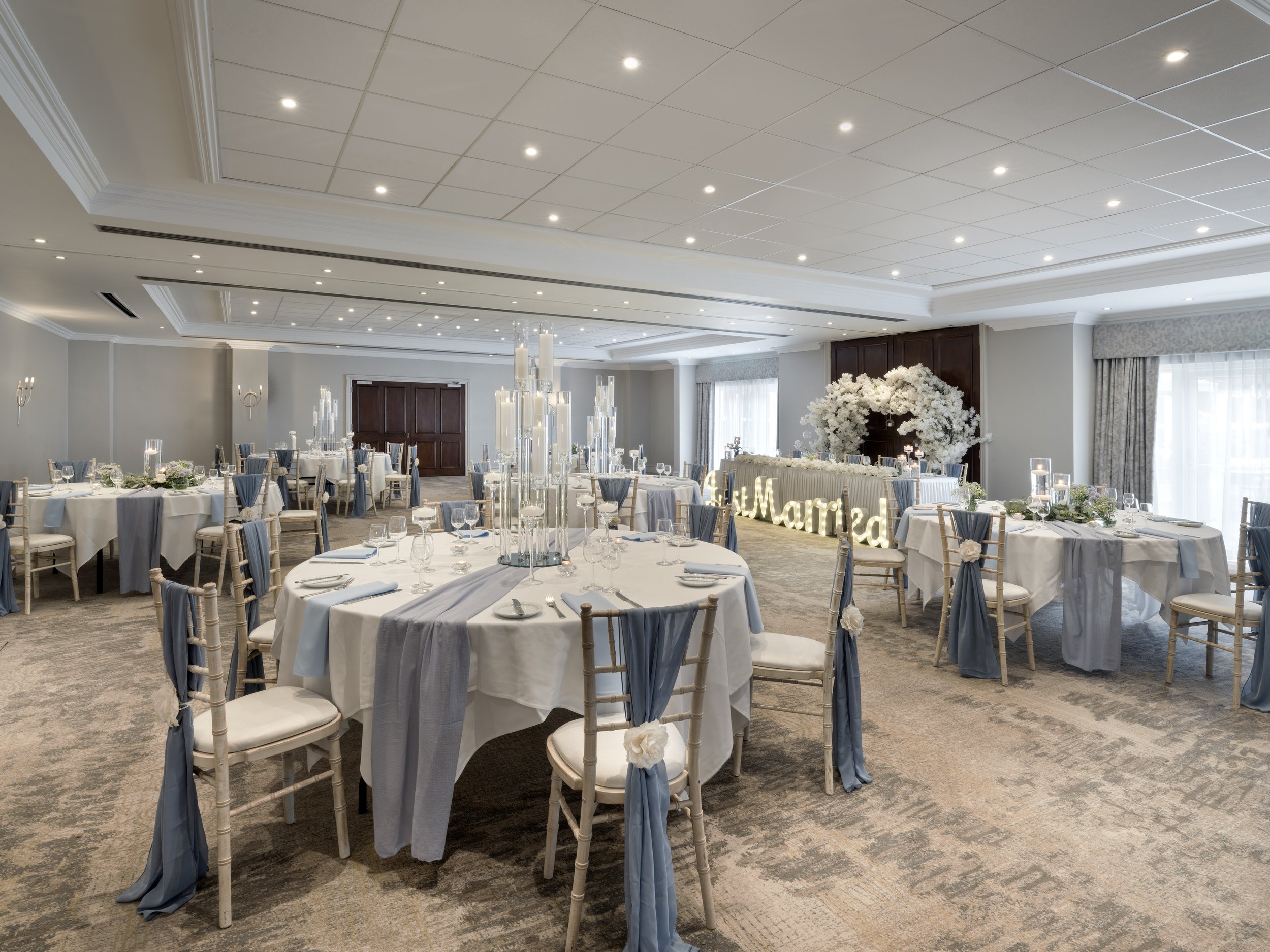 A banquet hall decorated for a wedding reception with round tables draped in white and blue tablecloths, elegant white chairs with blue sashes, and tall floral centerpieces. There is a wedding arch decorated with white flowers and an illuminated sign that says 'Married' in the background. The ceiling is fitted with multiple recessed lights, and large windows with curtains let in natural light.