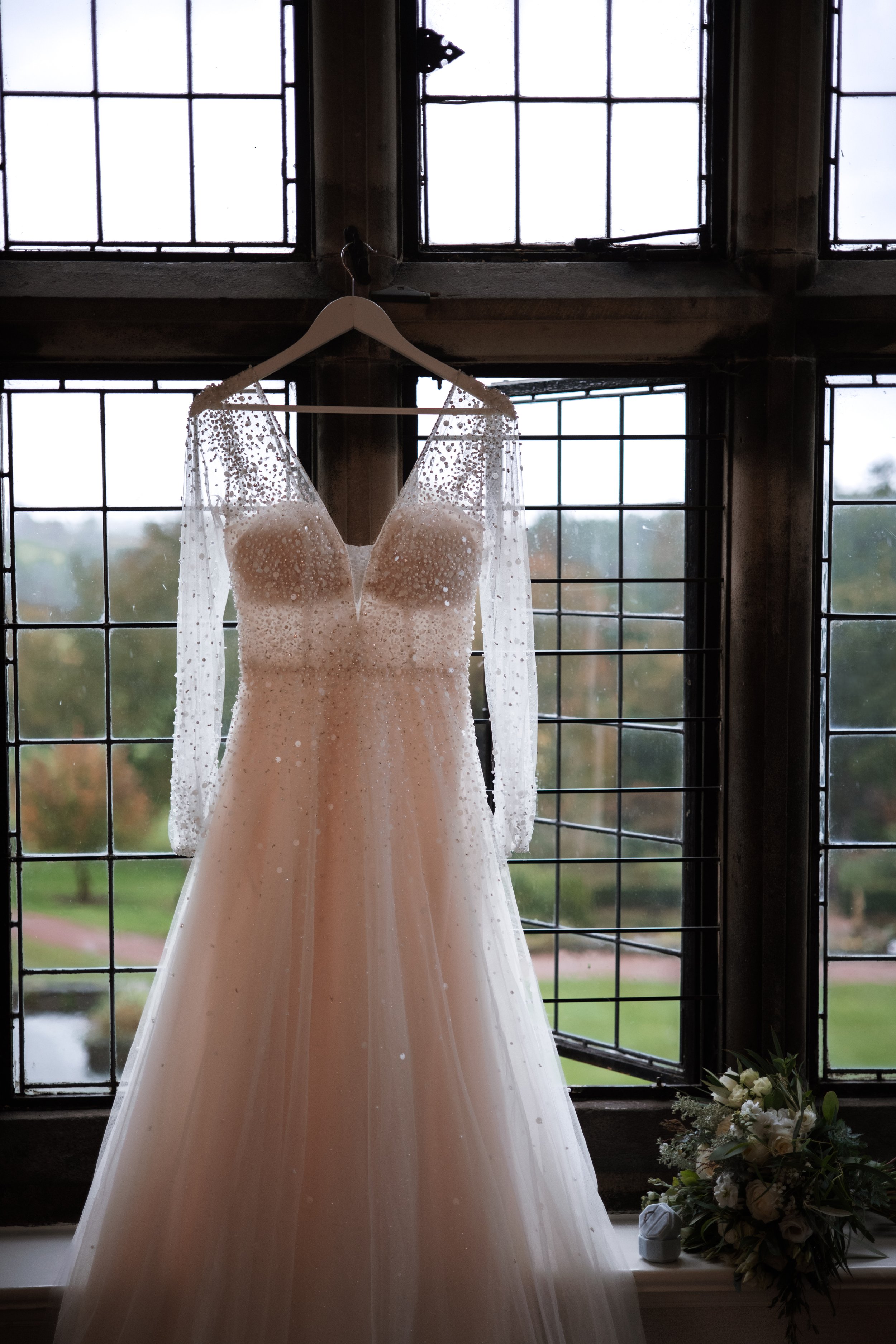 A wedding dress hanging in front of a large window with metal framework. The dress has a fitted bodice with a V-neckline and long sheer sleeves with small embellishments, and a flowy skirt. A bouquet of white flowers and greenery rests on the windowsill.