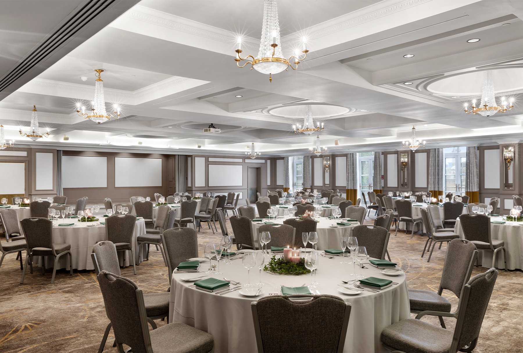 Large banquet hall decorated for an event with round tables covered in white tablecloths, set with glasses, plates, and cloth napkins, surrounded by gray upholstered chairs, adorned with candles and greenery centerpieces, illuminated by chandeliers and natural light from windows.