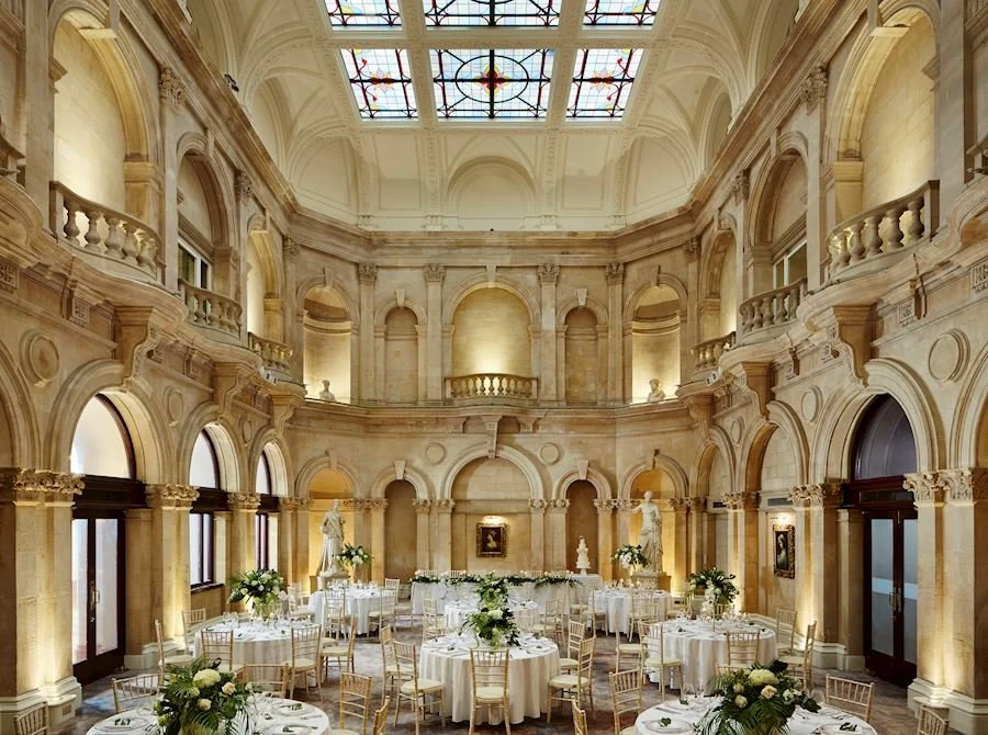 Elegant banquet hall with high arched ceilings, stained glass skylight, and classical statues, decorated with white tablecloths, floral centerpieces, and arranged for a formal event.