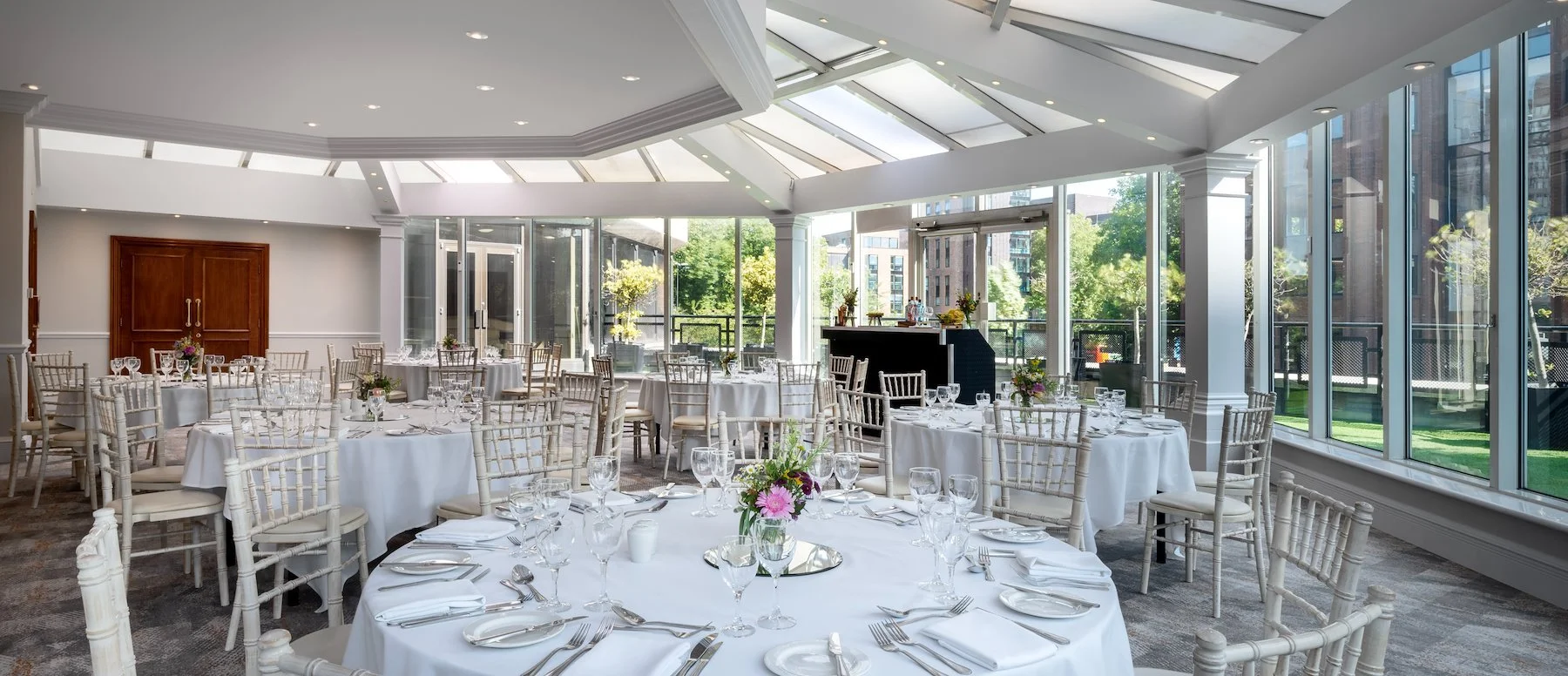 Elegant banquet room with round tables covered in white tablecloths, set with glassware, silverware, and floral centerpieces. Large windows and skylights let in natural light, with views of greenery and city buildings outside.