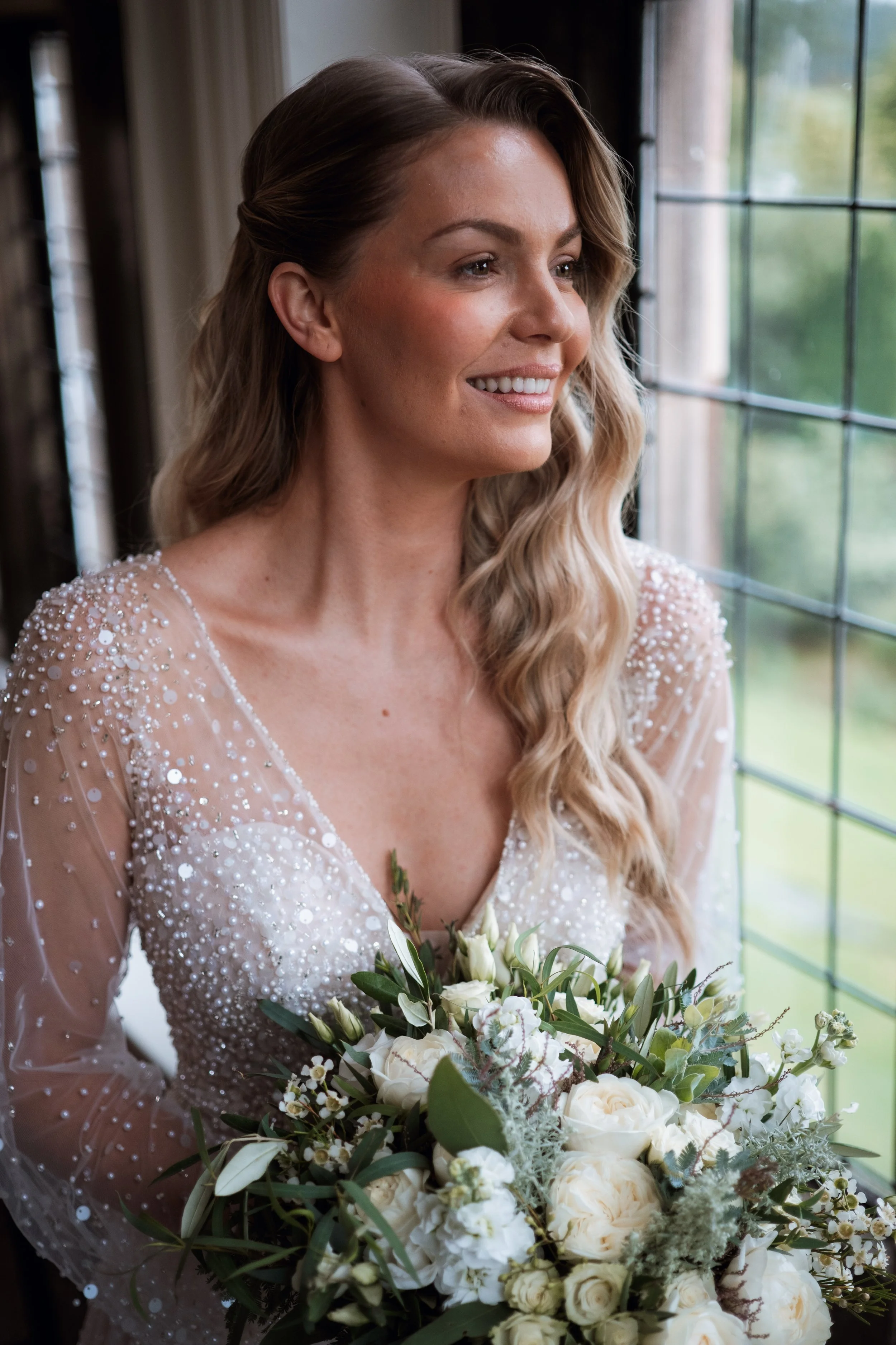 A smiling woman in a wedding dress holding a bouquet of white flowers near a window with a green outdoor view.