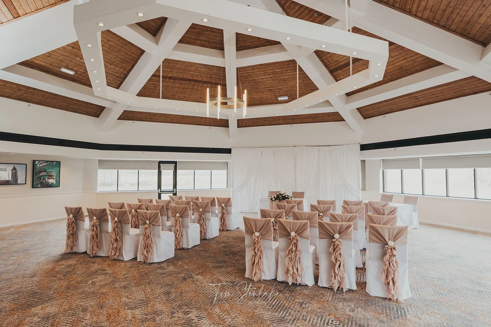 Elegant indoor wedding ceremony setup with chairs decorated with peach-colored fabric, a white backdrop curtain, and a floral arrangement on a table in front.