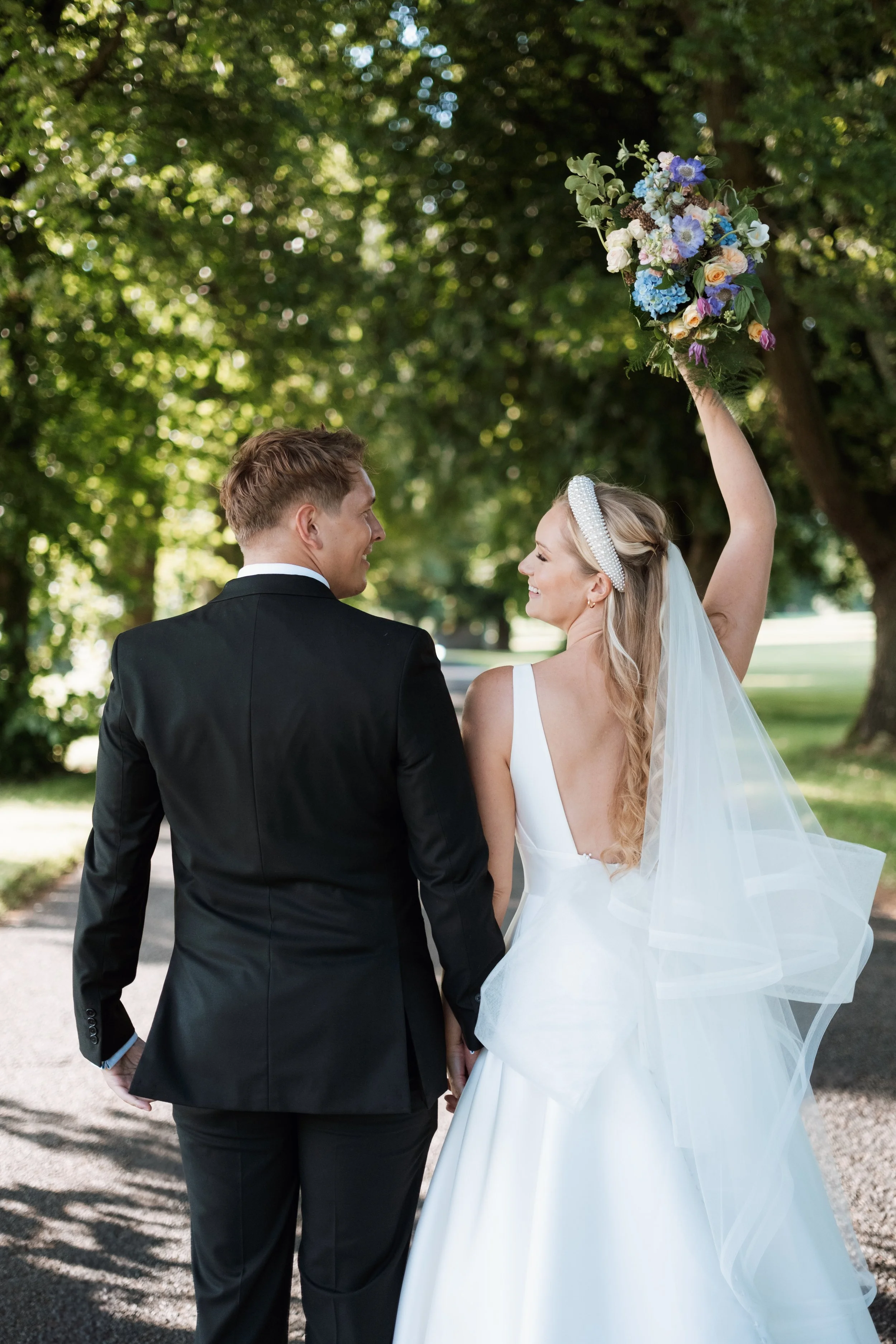 A bride and groom hold hands outdoors in a park, with the bride raising a bouquet of flowers above her head as they smile at each other.