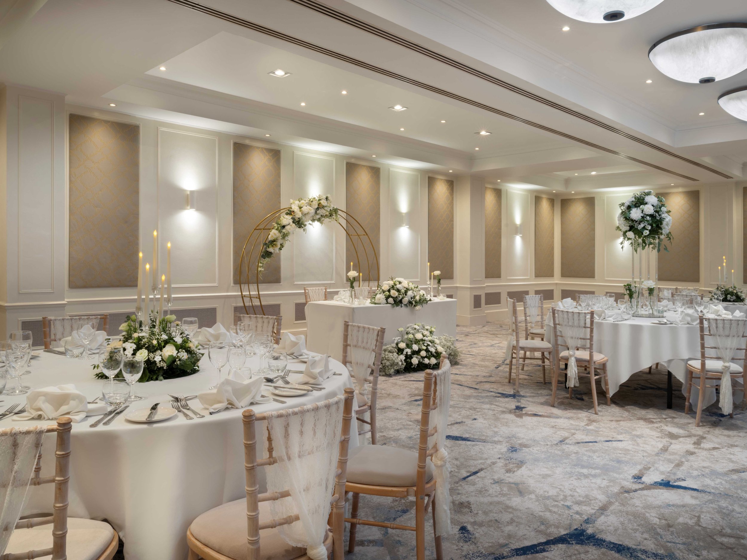 Elegant banquet hall decorated for a wedding reception with round tables, white tablecloths, floral centerpieces, and gold accents, featuring a floral arched backdrop and soft lighting.