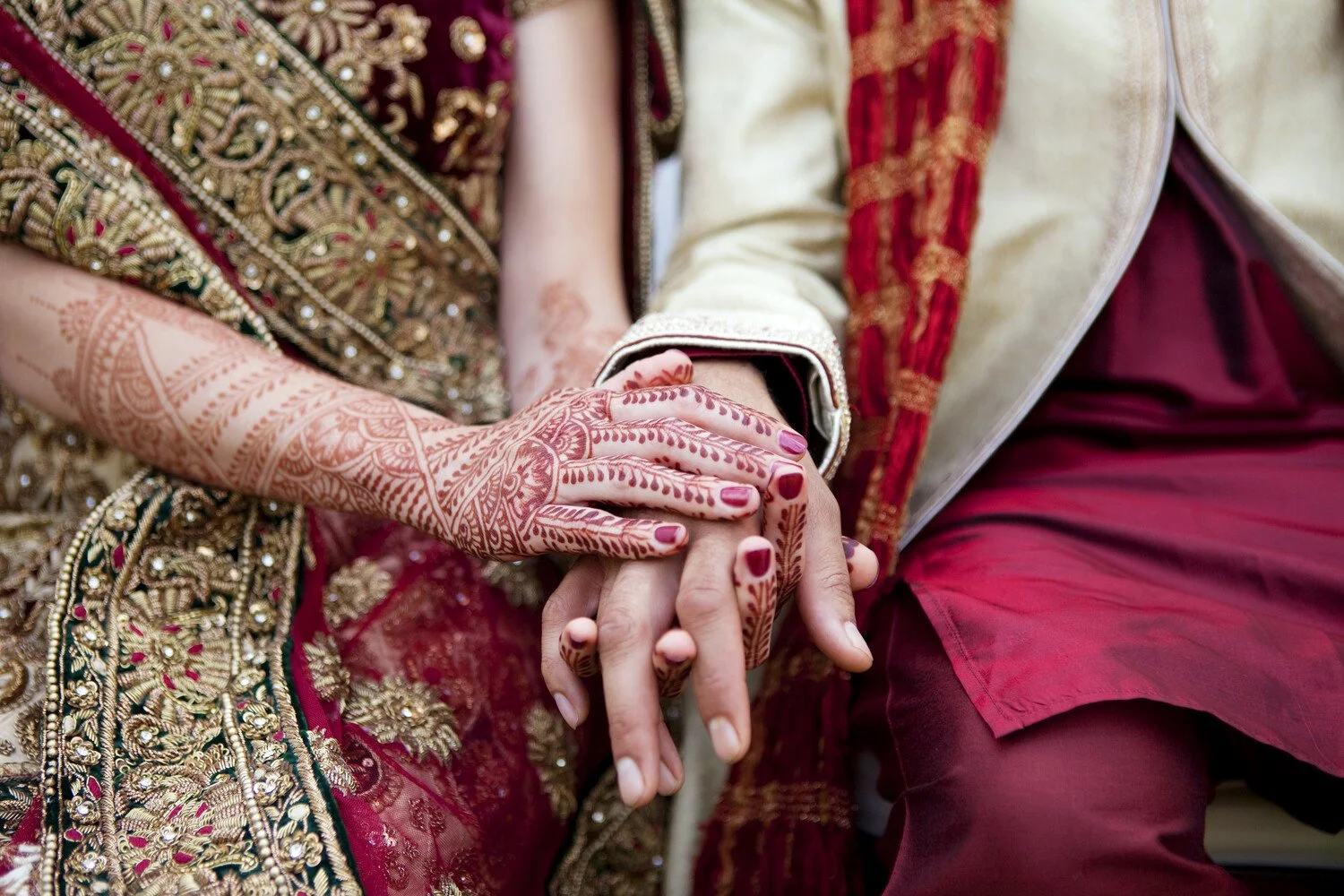 A couple holding hands, dressed in traditional Indian wedding attire with intricate embroidery and henna designs on the woman's hands.