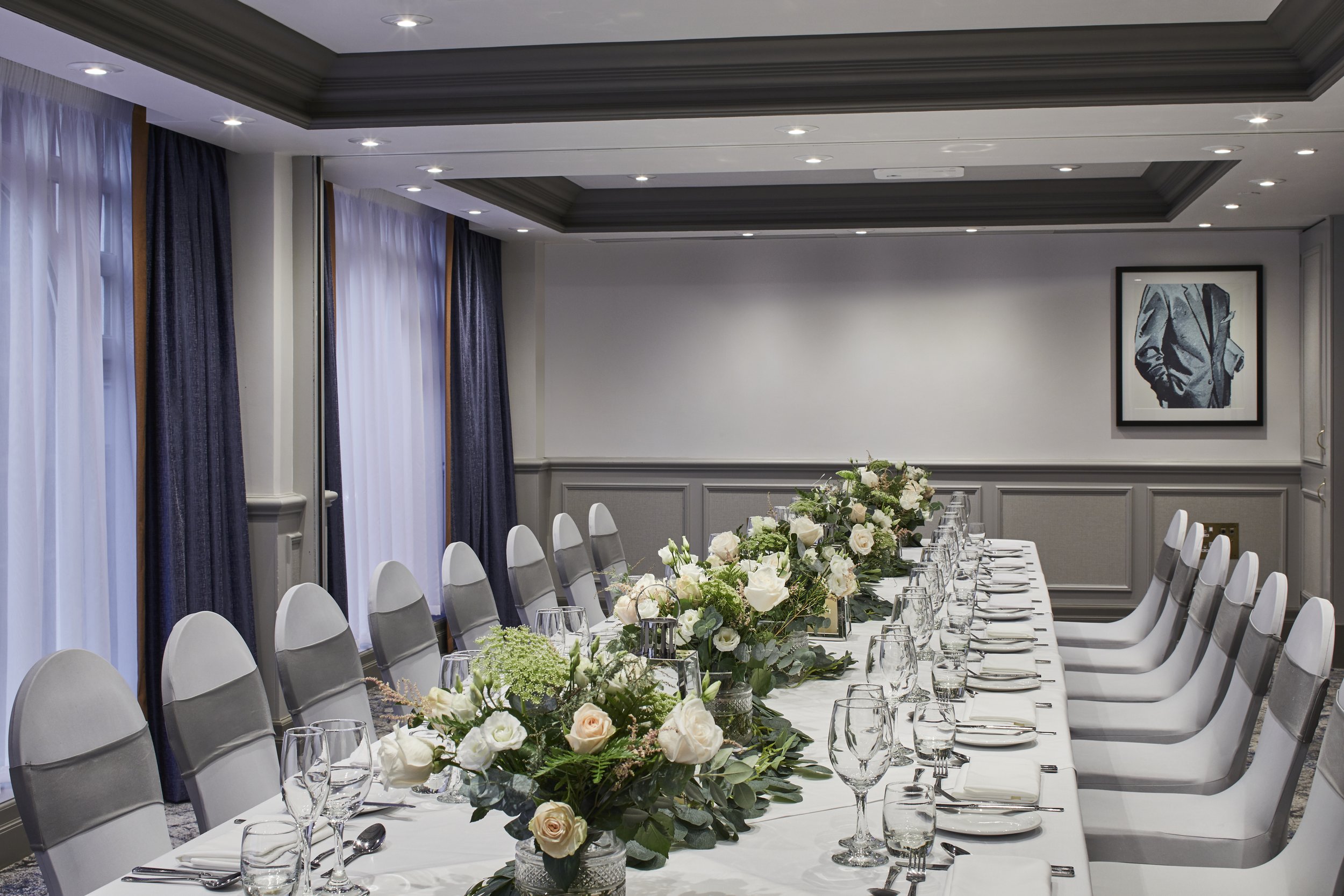 Elegant dining room set for a formal event with a long table decorated with white and blush roses, white tablecloth, glassware, and silverware, surrounded by white chairs, with large windows draped with white and dark curtains, and a framed black-and-white picture of a man in a suit on the wall.
