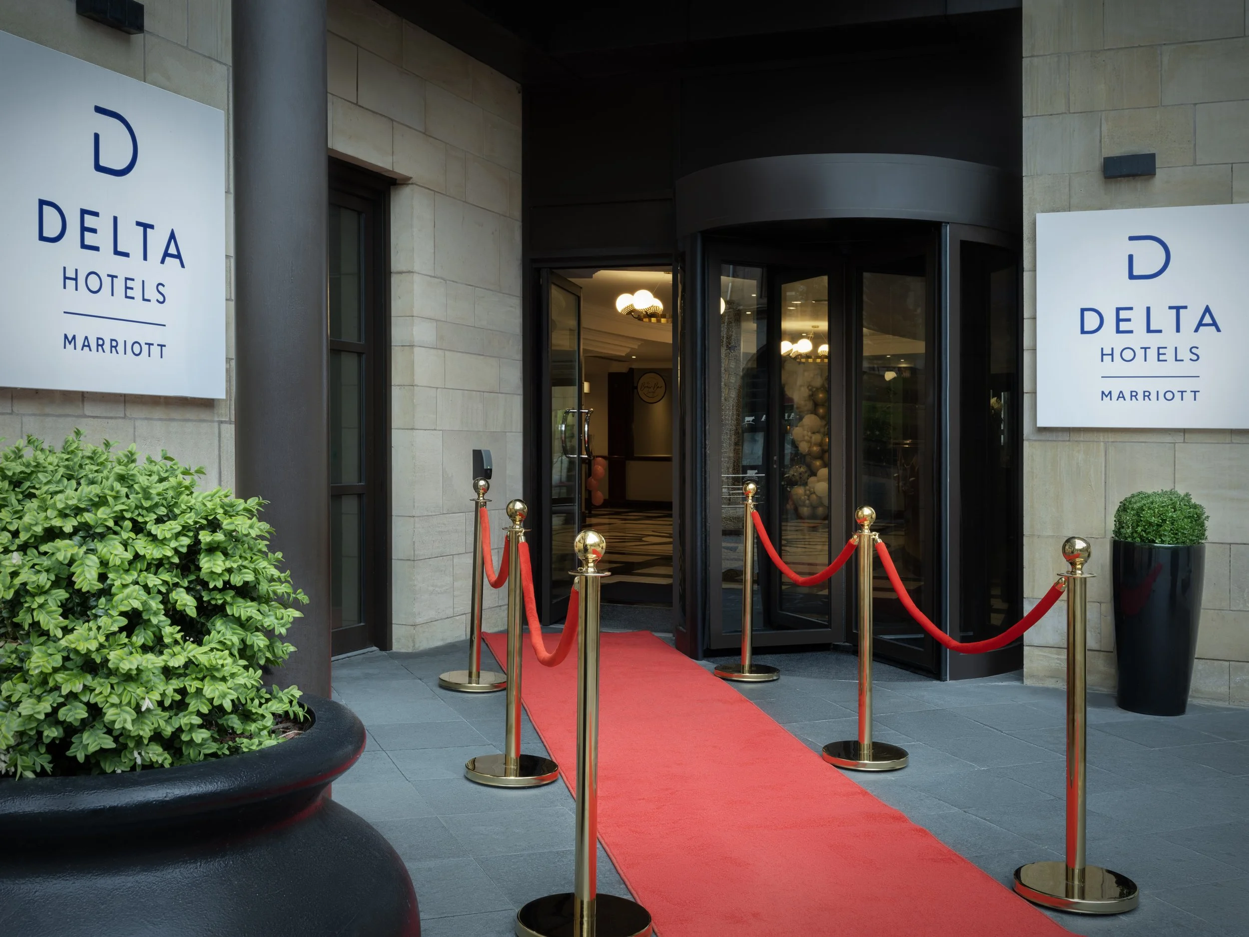 Red carpet walkway with gold stanchions and red velvet ropes leading to the entrance of Delta Hotels by Marriott, with two large white signs displaying the hotel logo on either side and greenery in black planters.