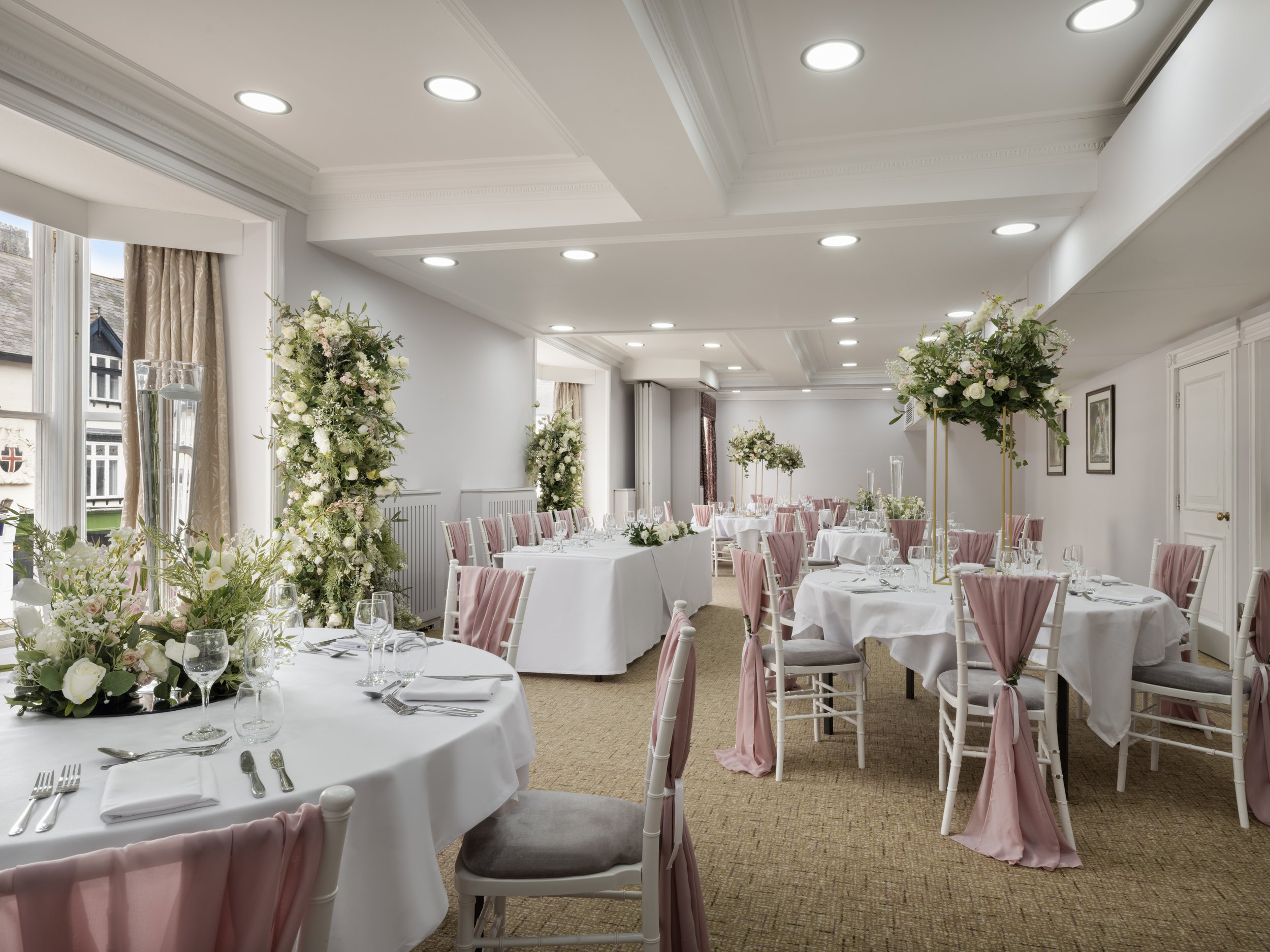 An elegantly decorated banquet hall with round tables draped in white tablecloths, pink chair sashes, and floral centerpieces. The room has large windows with beige curtains, white walls, and recessed ceiling lights.