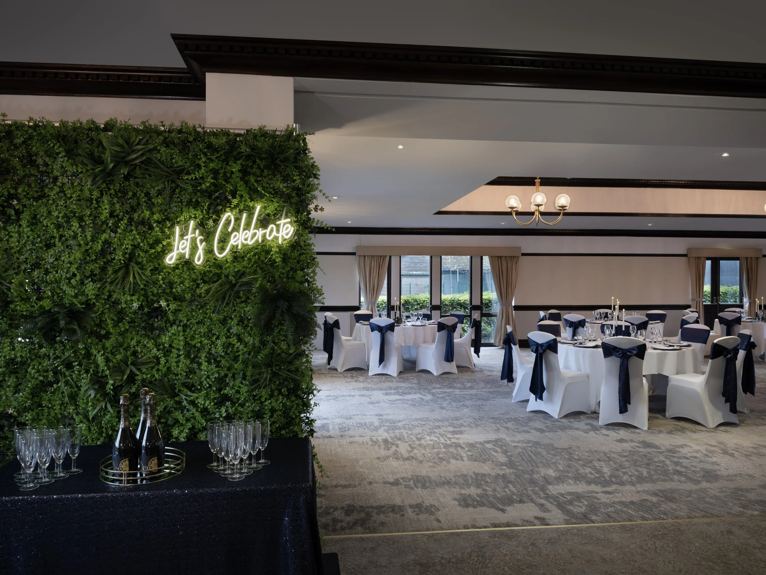 An event space decorated for celebration with a green leafy wall and a neon sign saying 'Let's Celebrate', set up with round tables and white chairs with navy ribbons, large windows, beige curtains, and a chandelier.