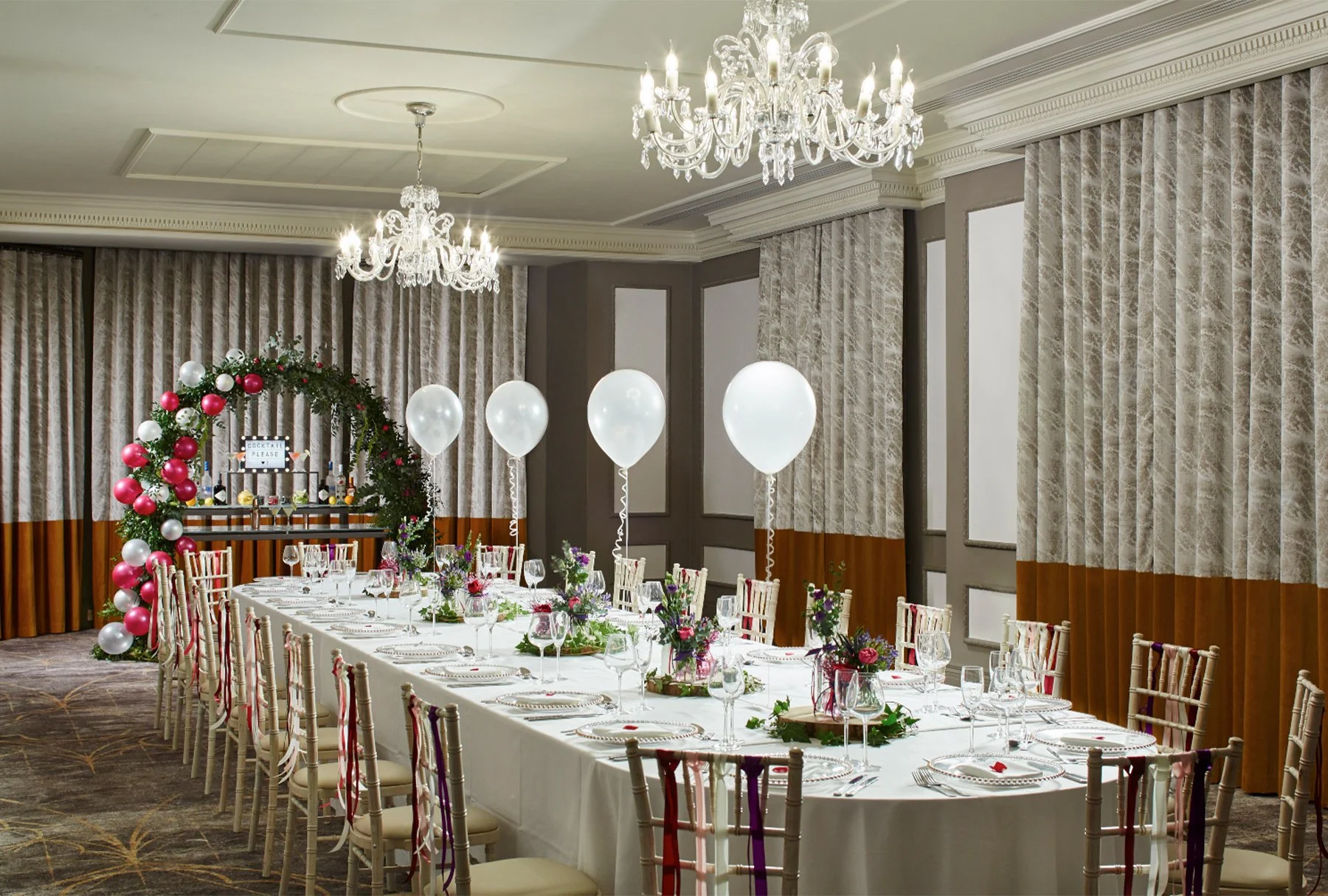 Elegant banquet hall decorated for a celebration with a long table covered in a white tablecloth, adorned with flower arrangements, glasses, and plates. There are large white balloons along the table. In the background, a festive arch decorated with red and white ornaments and green foliage holds bottles and a sign. The room features chandeliers, ornate molding, and patterned curtains.