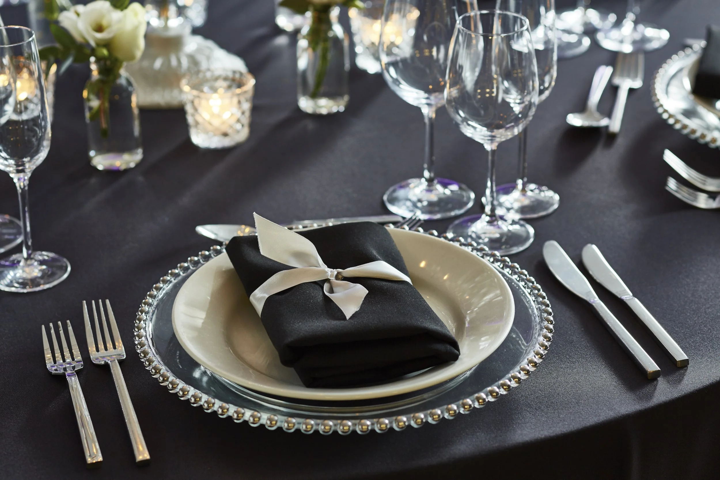Elegant dining table set with clear wine glasses, silverware, a black napkin with a white ribbon, and decorative candles on a dark tablecloth.