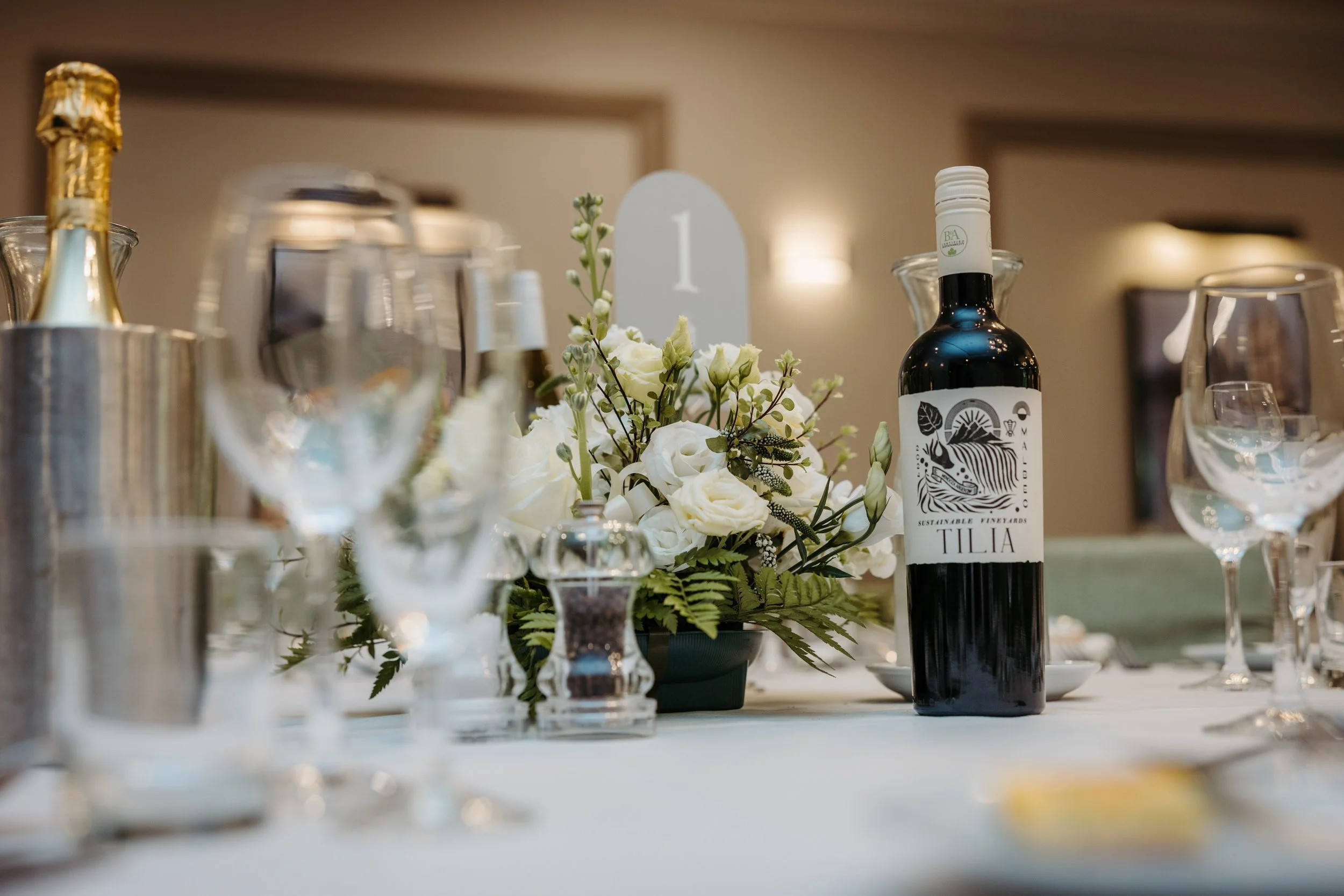 Table centerpiece with white flowers and a bottle of Tilia wine surrounded by glasses and silverware at an event or celebration.