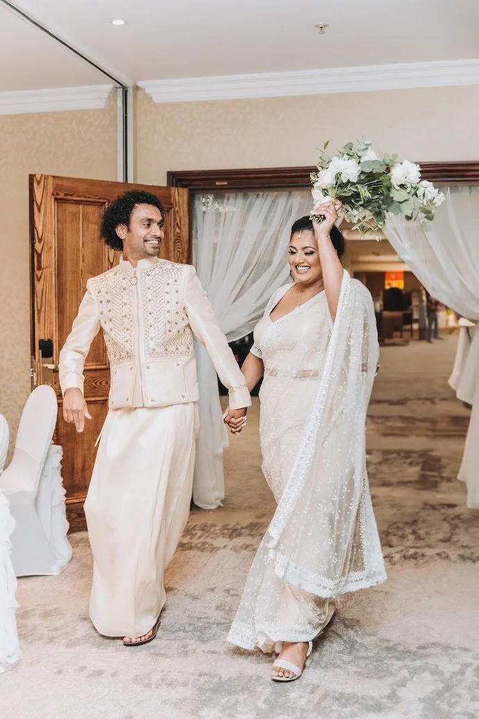 A bride and groom dance at their wedding reception, holding hands and smiling. The bride is lifting a bouquet of white flowers and greenery. They are dressed in traditional white attire, with the bride wearing a saree or lehenga and the groom in a cream-colored sherwani. The setting is decorated with light-colored drapes and a wooden door in the background.
