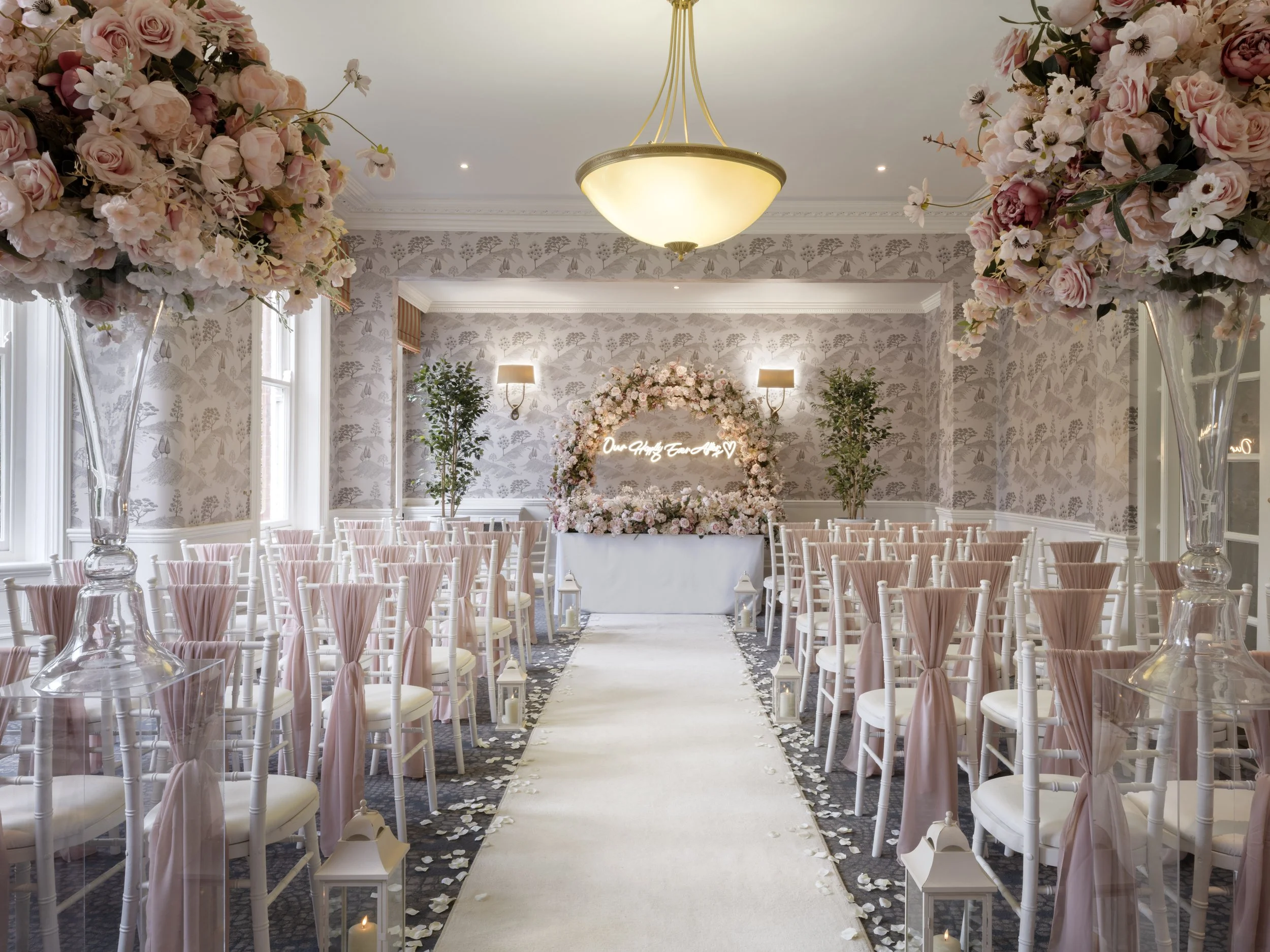 A decorated wedding or event venue with floral arrangements, a white carpet aisle, lanterns with candles, and seating chairs with pink sashes.