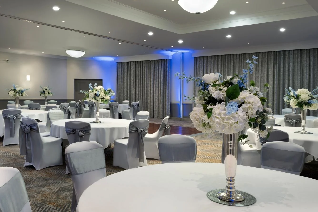 Elegant event space decorated with white tablecloths, silver chair sashes, and floral centerpieces in tall vases, set up for a banquet or wedding reception.