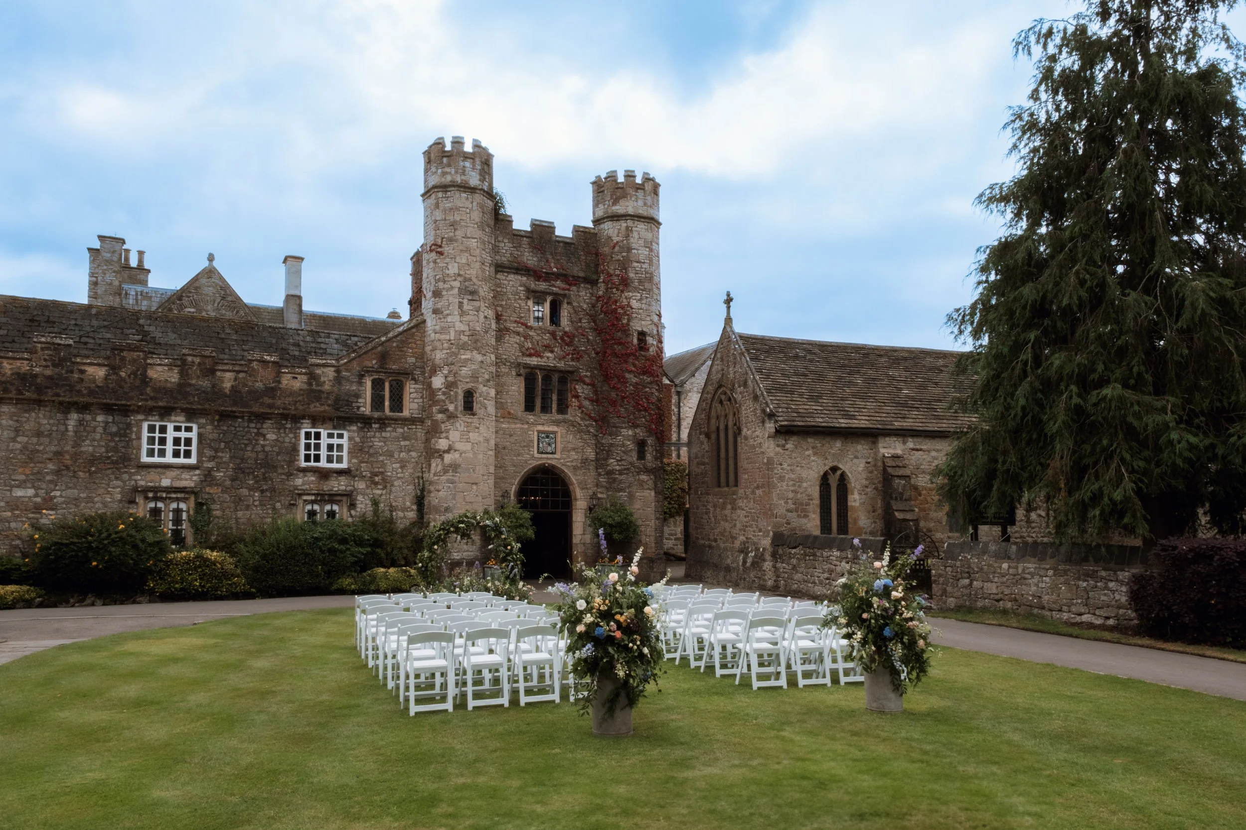 A historic stone castle with turreted towers in the background, decorated with red climbing plants, and a wedding setup in the foreground with rows of white chairs and floral arrangements on tall vases, set on a well-manicured lawn under a partly cloudy sky.