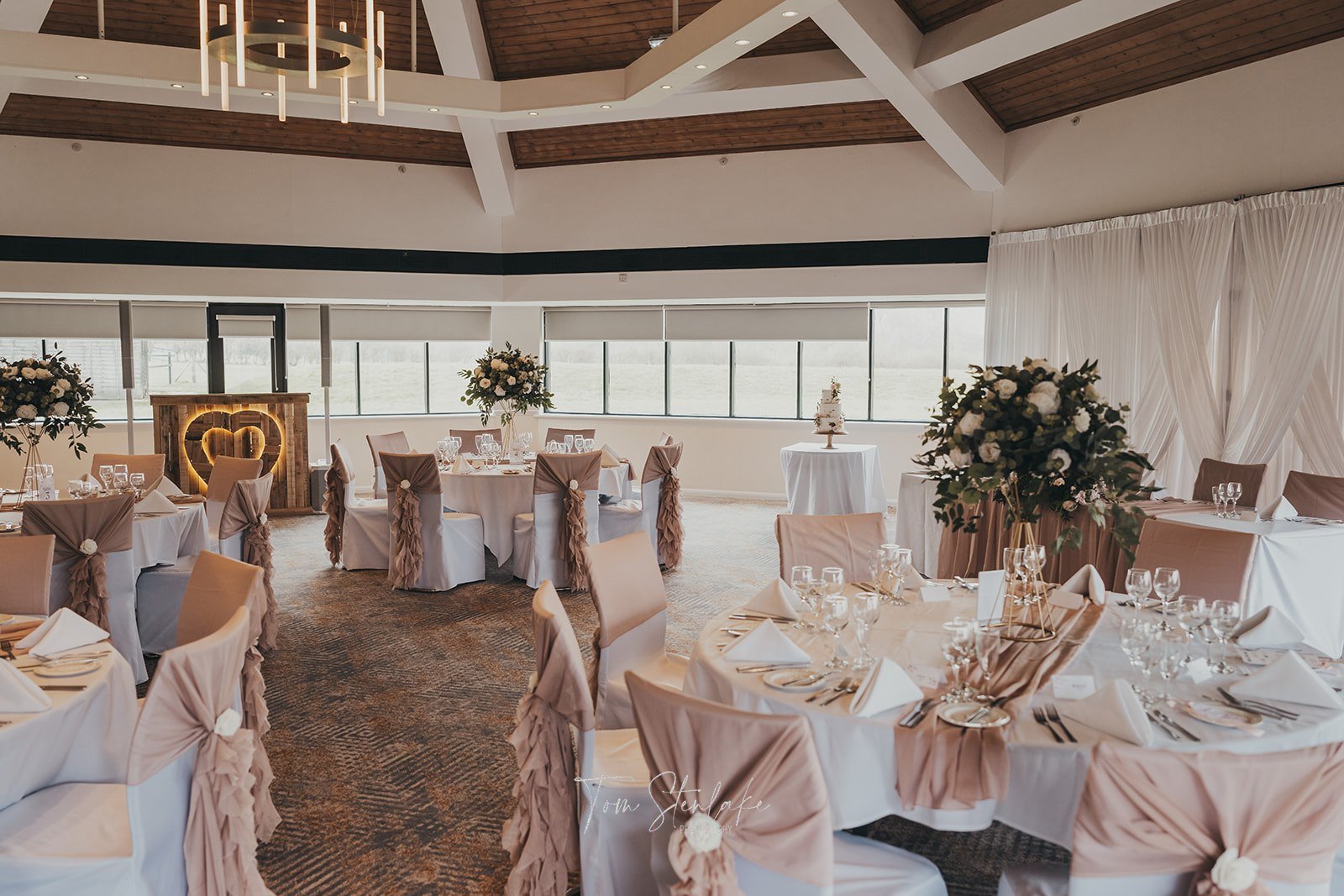 Wedding reception hall with round tables decorated with white tablecloths, beige chair covers with ruffled sashes, floral centerpieces, and a cake on a separate table. Large windows, wooden ceiling, and a heart-shaped light installation in the background.