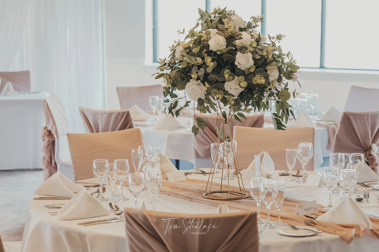 Elegant banquet table setup with a large floral centerpiece featuring white roses and greenery, surrounded by folded white napkins, wine glasses, and cutlery, in a bright room with large windows.