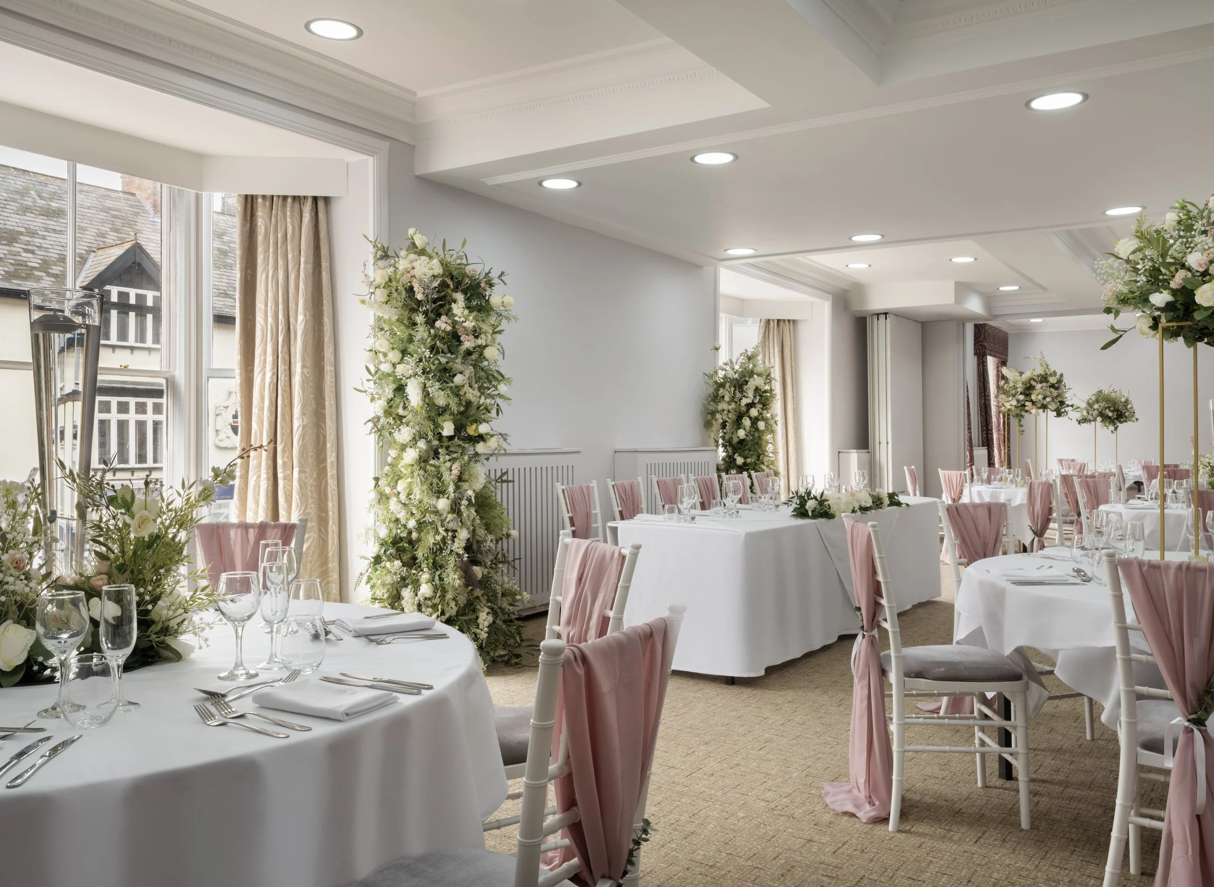 Elegant banquet hall decorated with tall floral arrangements, round tables with white tablecloths, pink chair sashes, glassware, and cutlery, large windows with beige curtains, and a ceiling with recessed lighting.