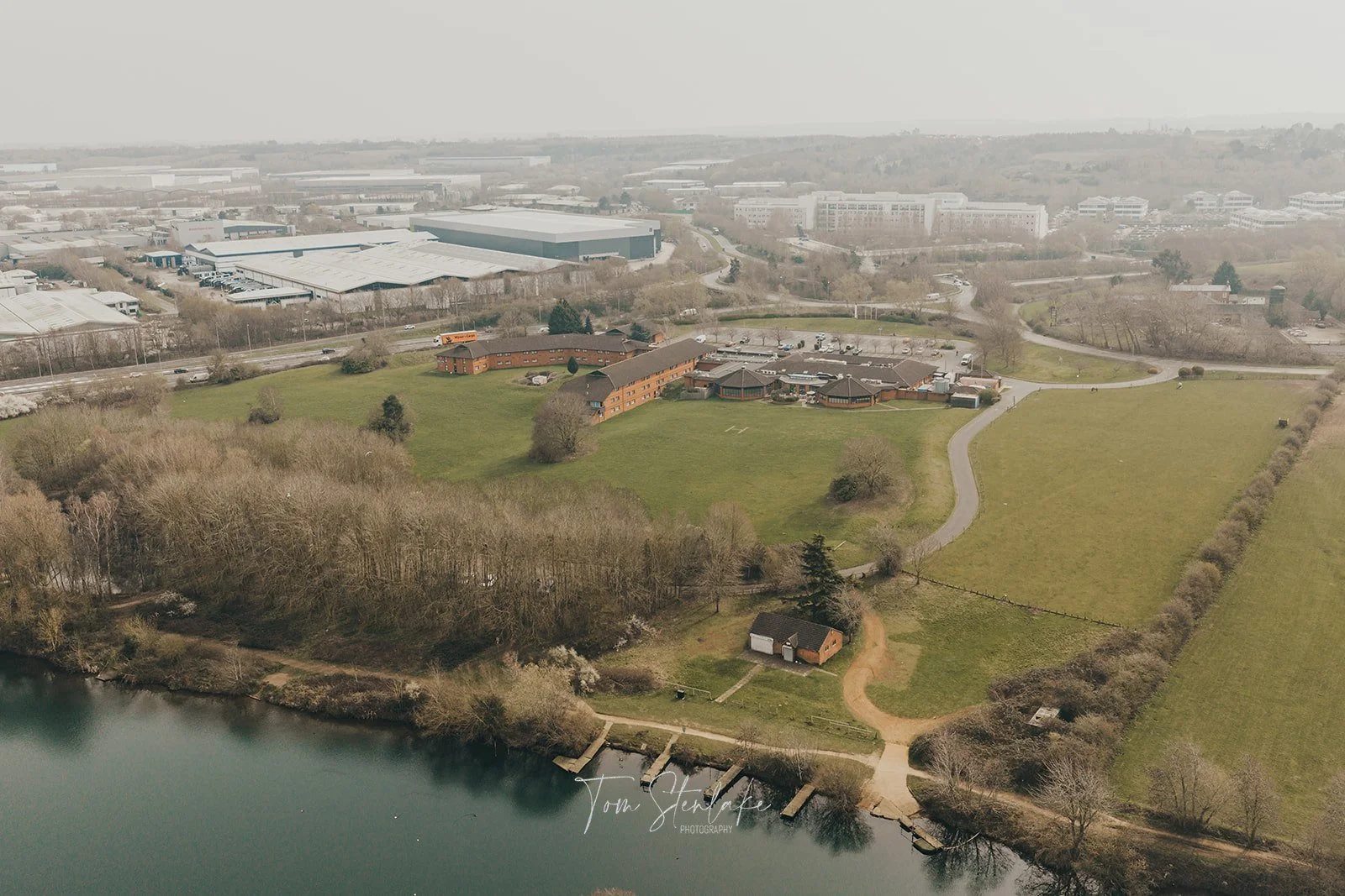 Aerial view of a landscape with a body of water in the foreground, grassy areas, and trees, with buildings and industrial structures in the background on a hazy day.