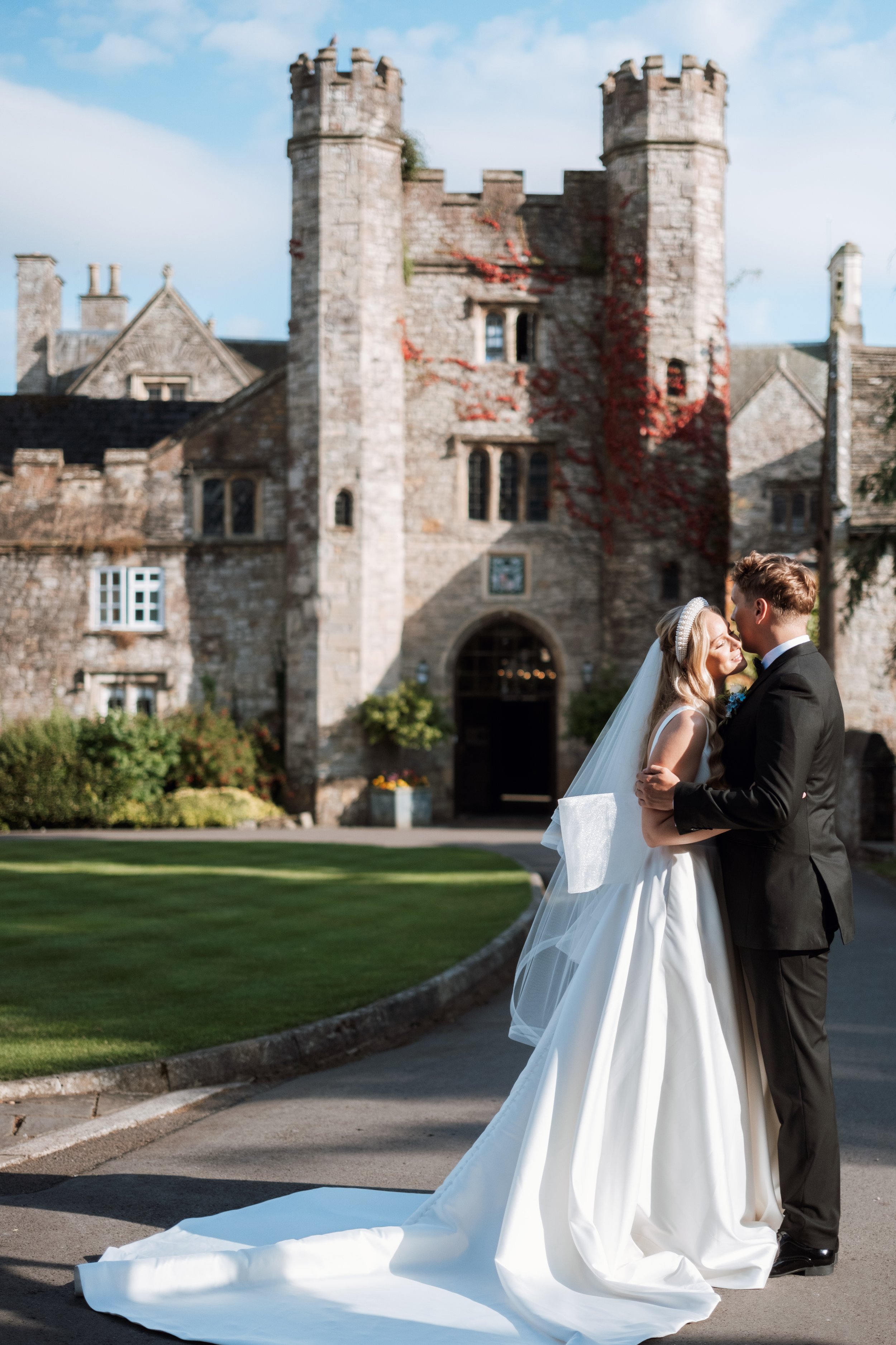 A bride and groom sharing a kiss outside a stone castle during their wedding, with the bride in a white gown and veil and the groom in a black suit, against the backdrop of a historic castle with towers and ivy, under a partly cloudy sky.