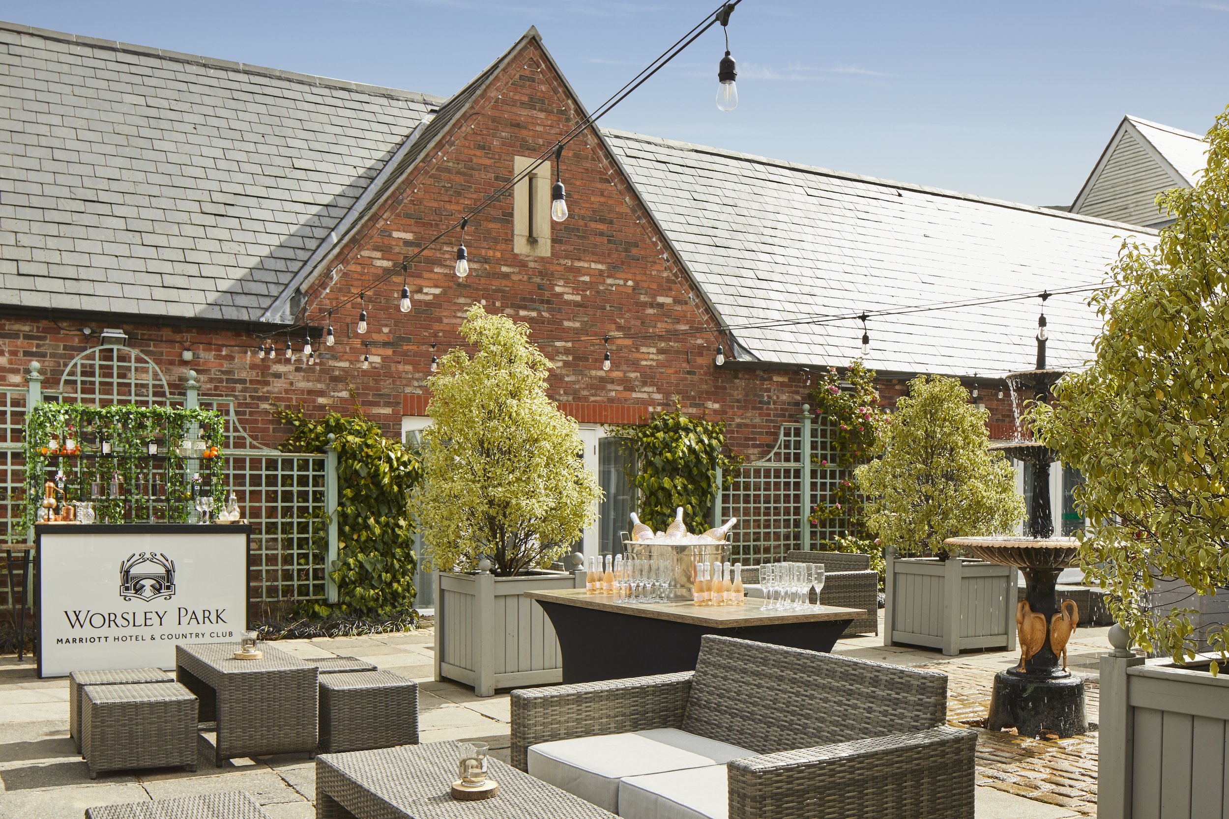 Outdoor patio area of Worsley Park Marriott Hotel & Country Club with string lights, tables with glasses, potted plants, and brick building with slate roof in the background.