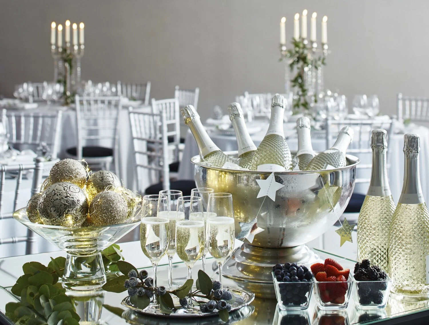 Elegant table setting with a silver ice bucket holding champagne bottles, decorated with star ornaments, in a decorated reception hall with white chairs and candle centerpieces.