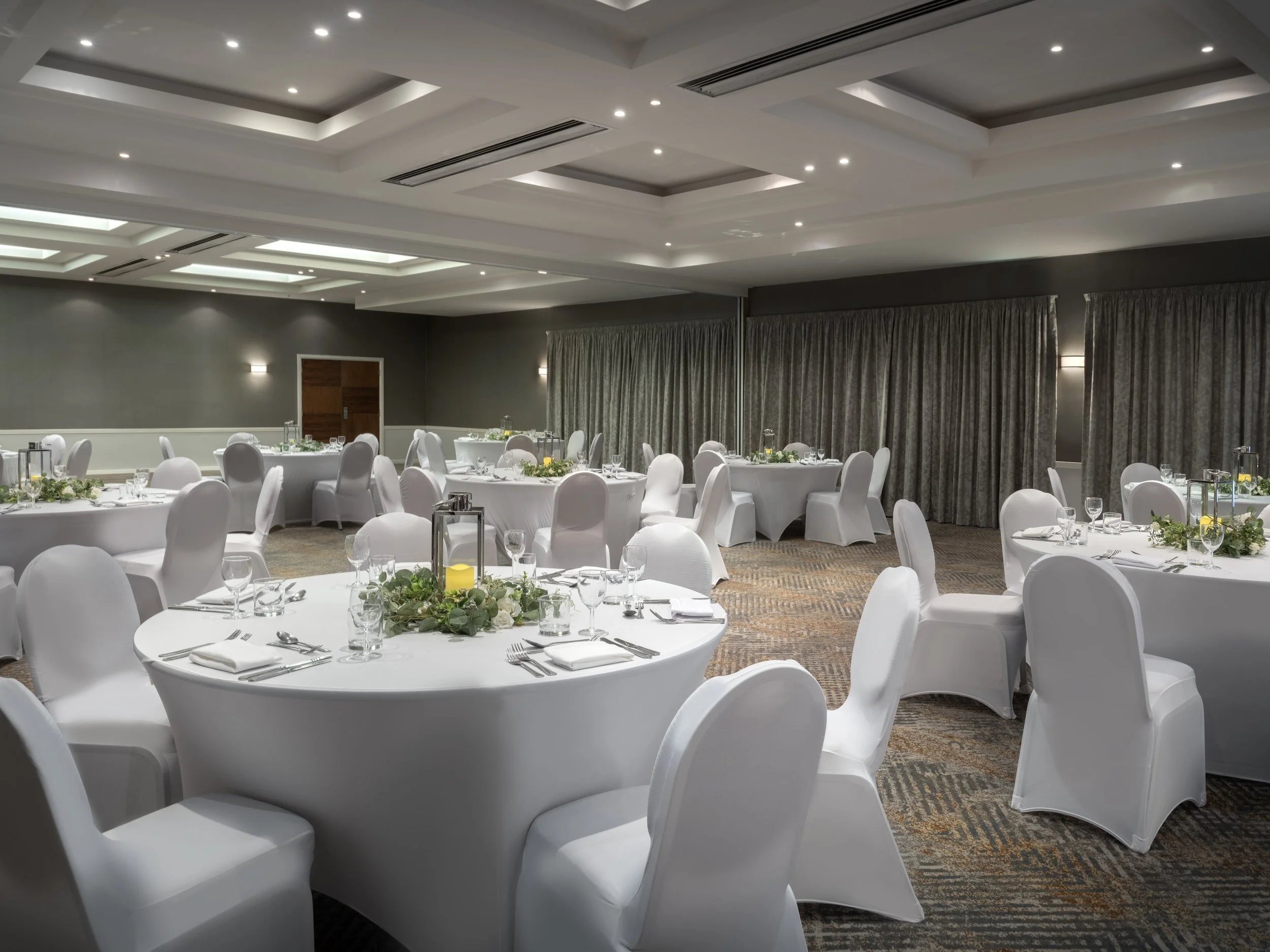 A banquet hall decorated for an event with round tables covered with white tablecloths and elegant place settings, including glasses and silverware. The centerpieces feature greenery and white flowers, and there are lanterns with candles on each table. The room has gray walls, heavy drapes, and a drop ceiling with recessed lighting.