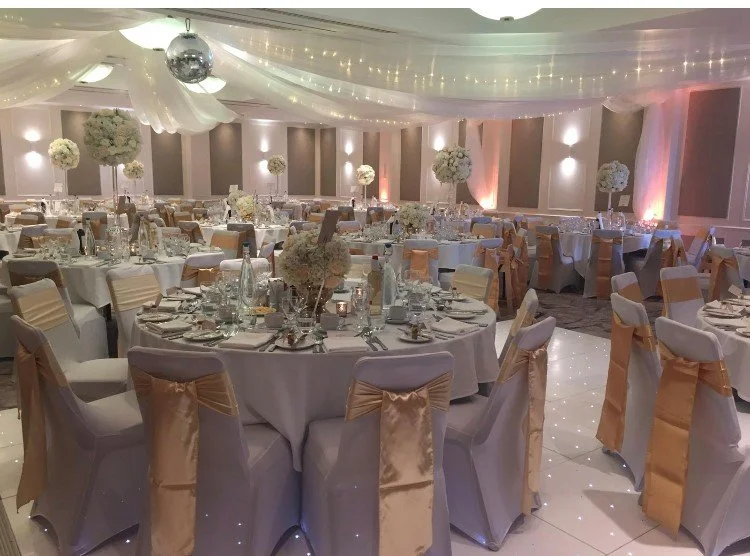 Elegant indoor wedding ceremony setup with rows of chairs decorated with green fabric and small white flowers, a white aisle runner, a floral arch at the altar, marble busts and framed artwork adorning the high stone walls.