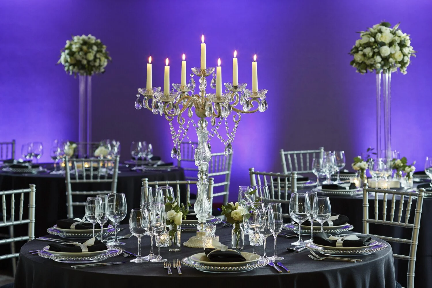 Elegant banquet table with black tablecloth, glassware, silverware, floral centerpieces, and a crystal chandelier candelabra against a purple-lit background.