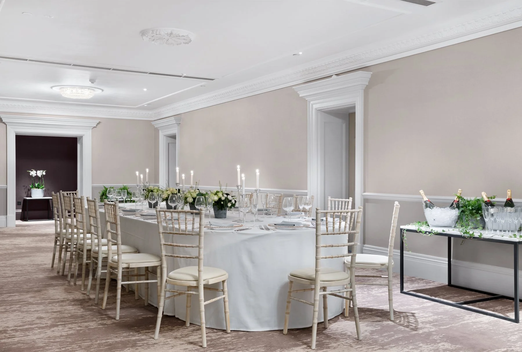 Elegant dining room decorated for a celebration with a long table set with white linens, chairs, wine glasses, and floral centerpieces, with a side table holding wine bottles and glasses, and a potted orchid in the background.